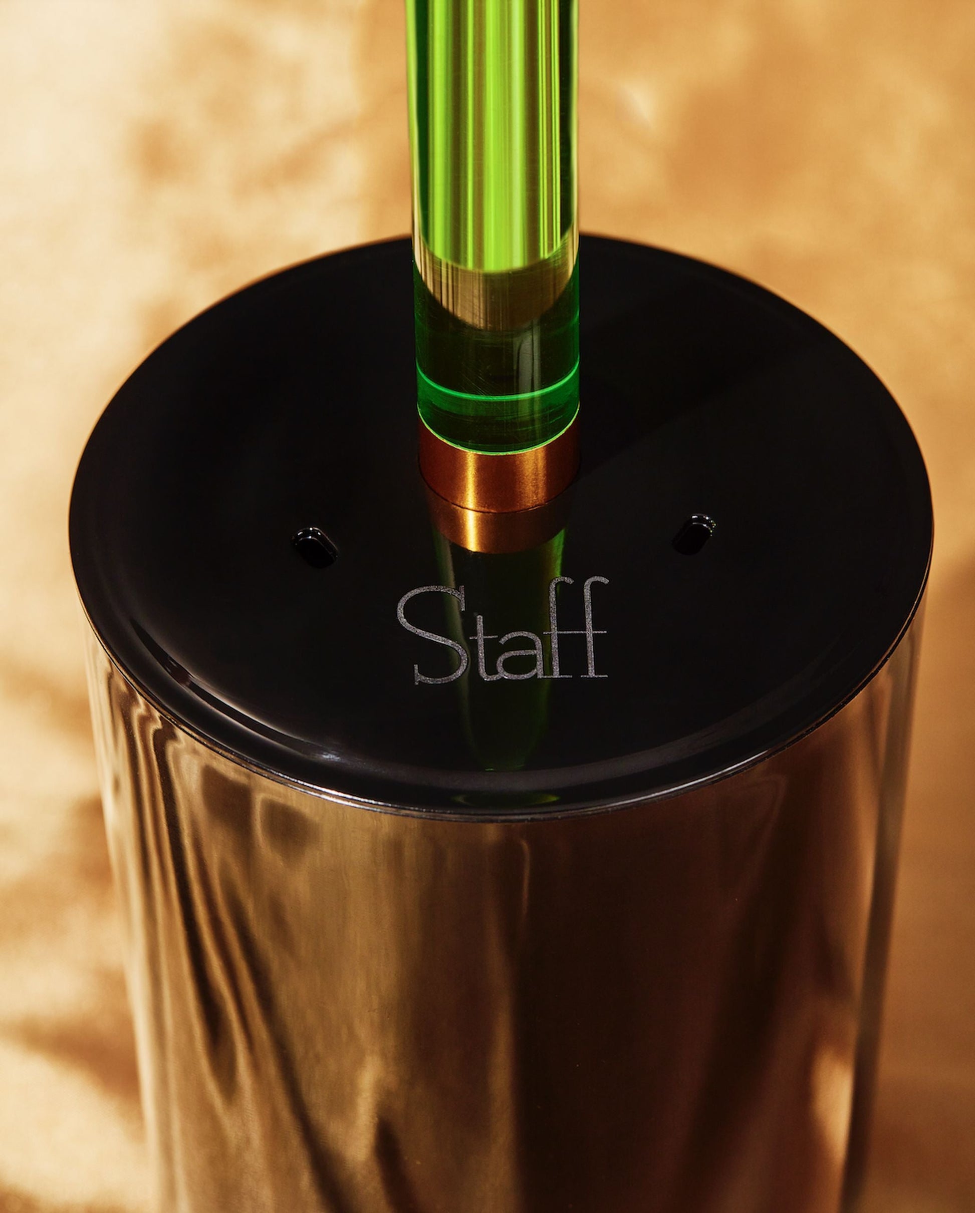 A close-up of the Staff The Toilet Brush - Green by Staff shows its shiny black base engraved with Staff and a translucent green acrylic handle, resembling a luxury toilet brush, set against a gold background.