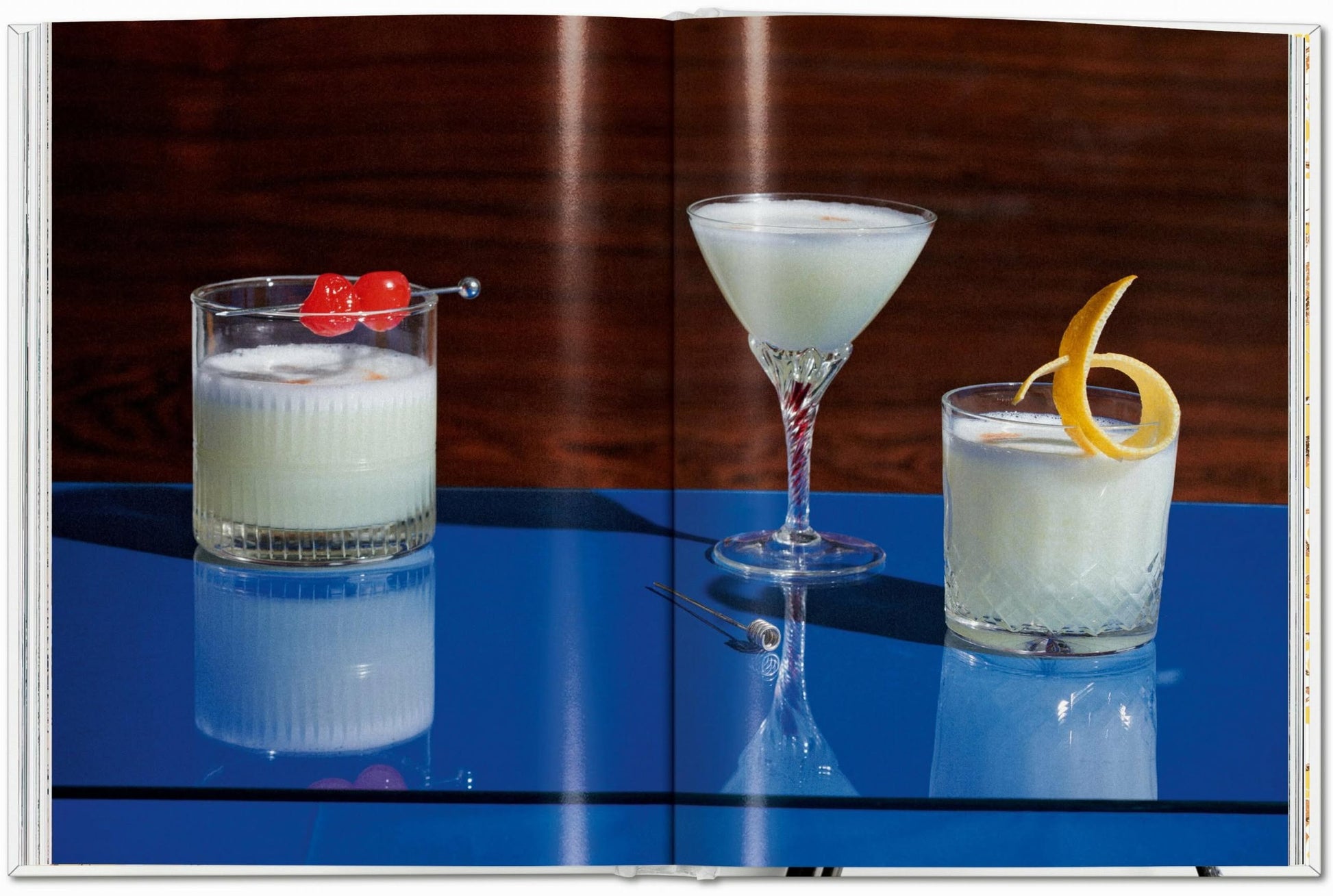 Three cocktails rest on a glossy blue surface beside a cocktail pick, capturing the spirit of TASCHEN The Gourmandʼs Egg: A Collection of Stories and Recipes—a blend where artful presentation and creative recipes meet.