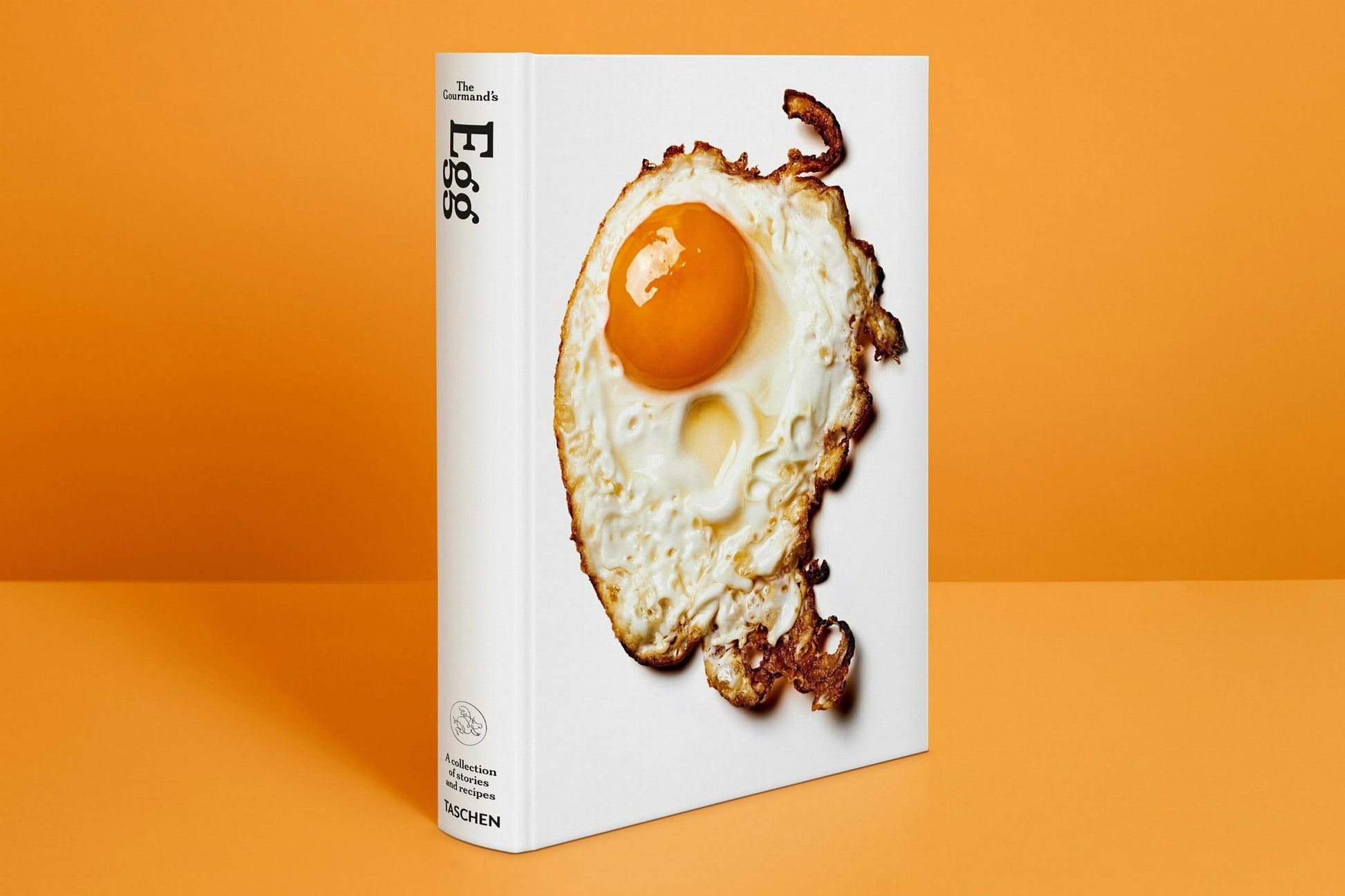 TASCHEN The Gourmands Egg: A Collection of Stories and Recipes stands upright against an orange backdrop, its cover showing a striking fried egg—celebrating food and creativity with unique recipes.