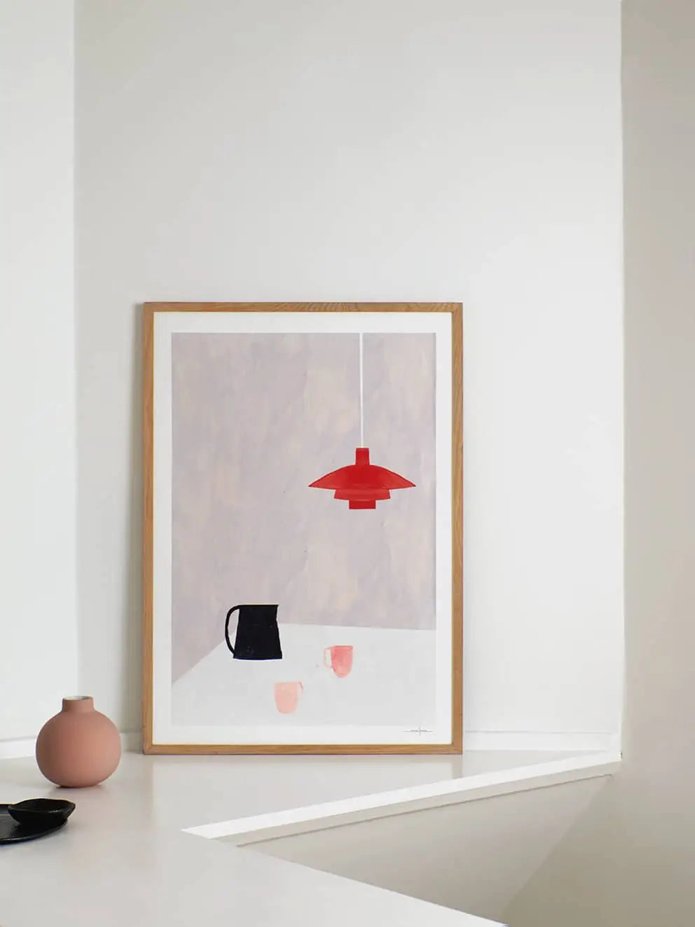 The Poster Clubs Ana Frois - Orange Pendant (50x70) print rests against a white wall, depicting a black pitcher, two pink cups, and an orange pendant lamp. A small pink vase and dark dish are placed nearby on the white surface.