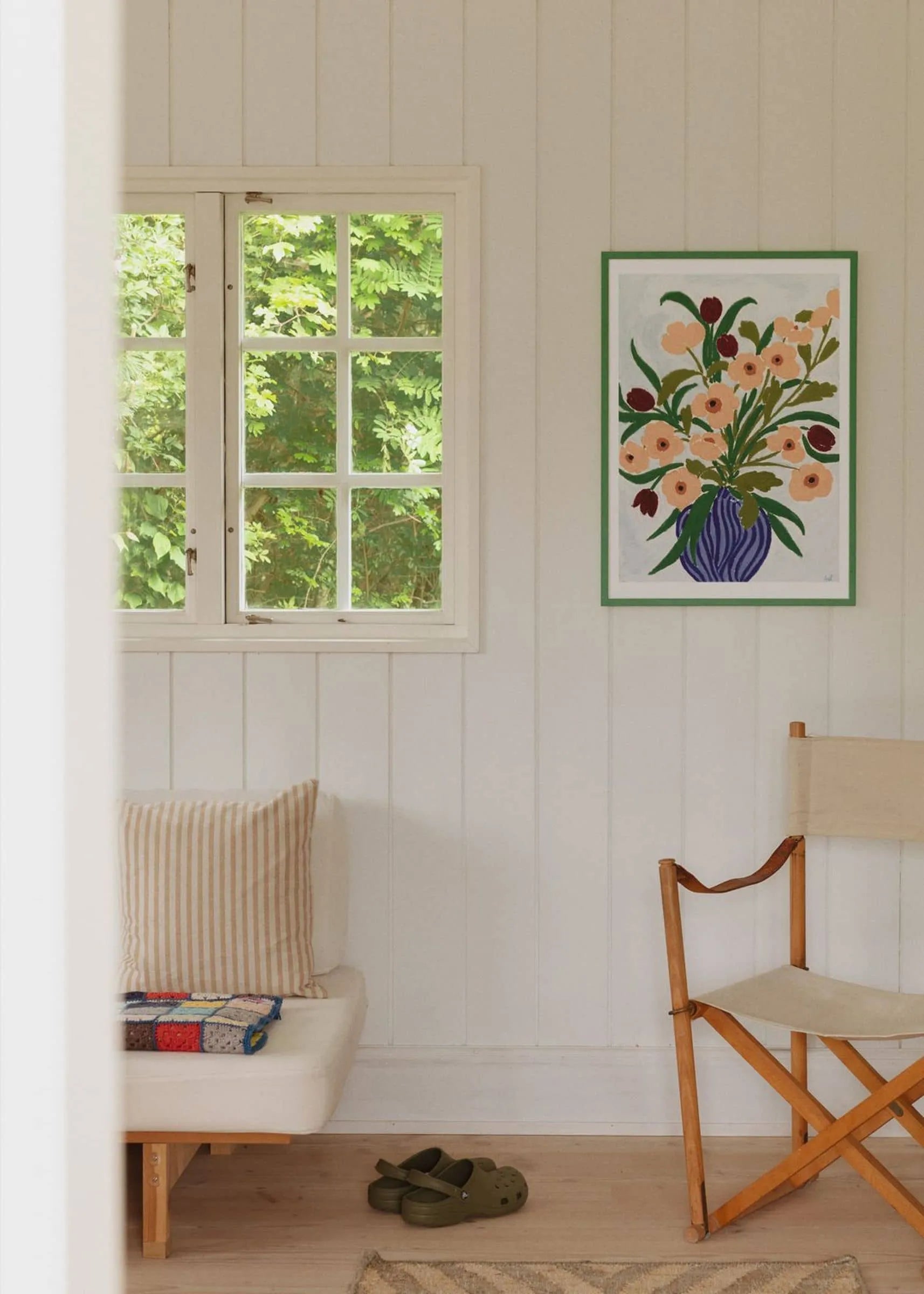 A bright room with green views, a white bench, striped pillows, books, wooden chair, green clogs, and Cerulean Still Life (30x40) by Anine Cecilie Iversen for The Poster Club displayed on the wall.