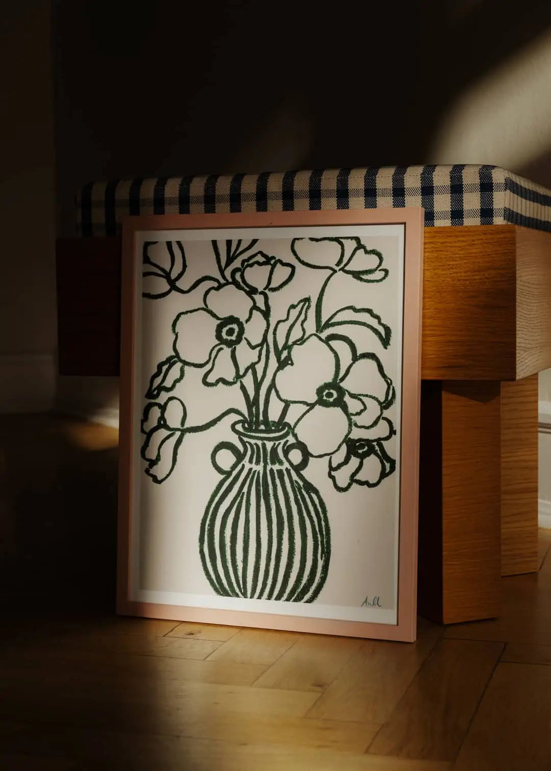 The Poster Club Ana Frois - Orange Pendant (50x70) features a fine art giclée print framed and bathed in sunlight, leaning against a bench with a blue and white checkered cushion on a wooden floor.