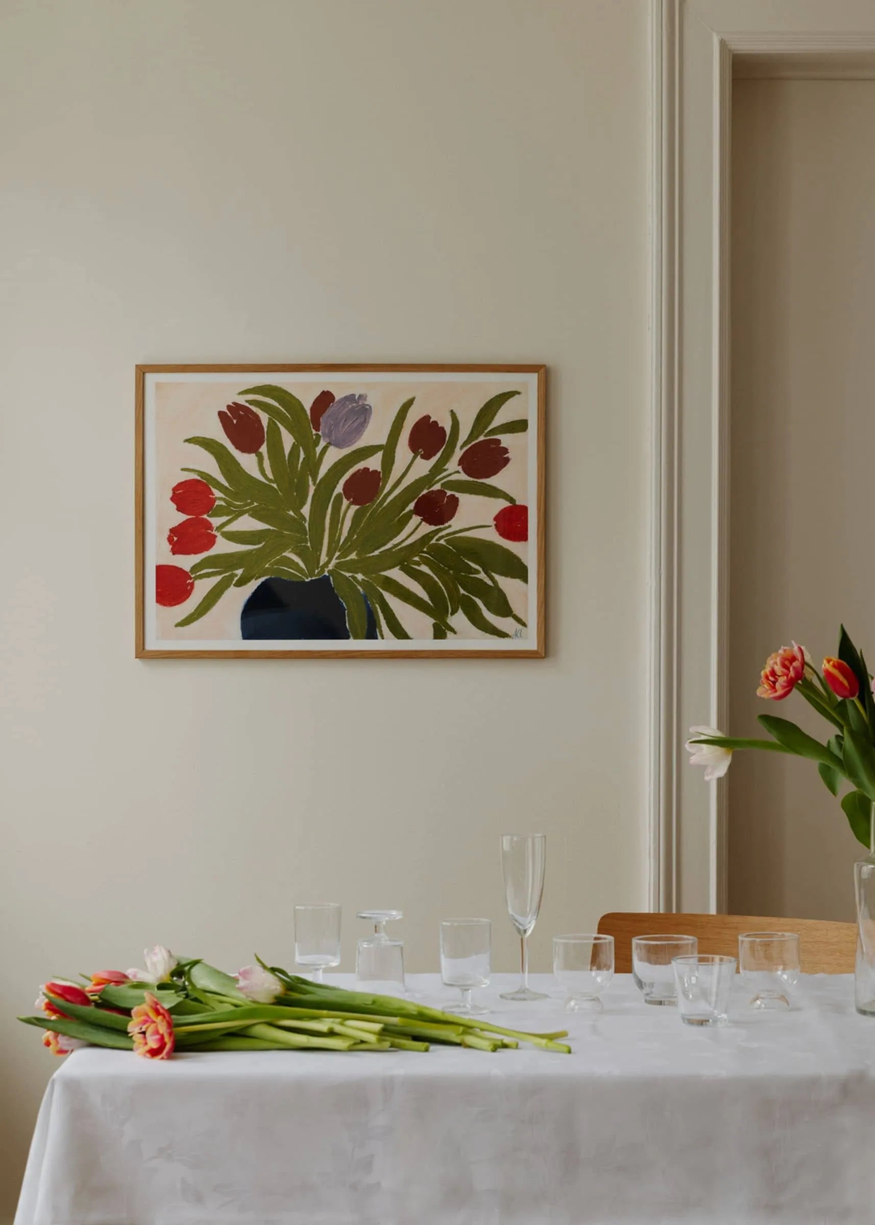 A table with empty glasses and tulips sits beneath Tulips in a Blue Vase (30x40) by Anine Cecilie Iversen for The Poster Club, featuring red tulips in a vase on a light-colored wall.