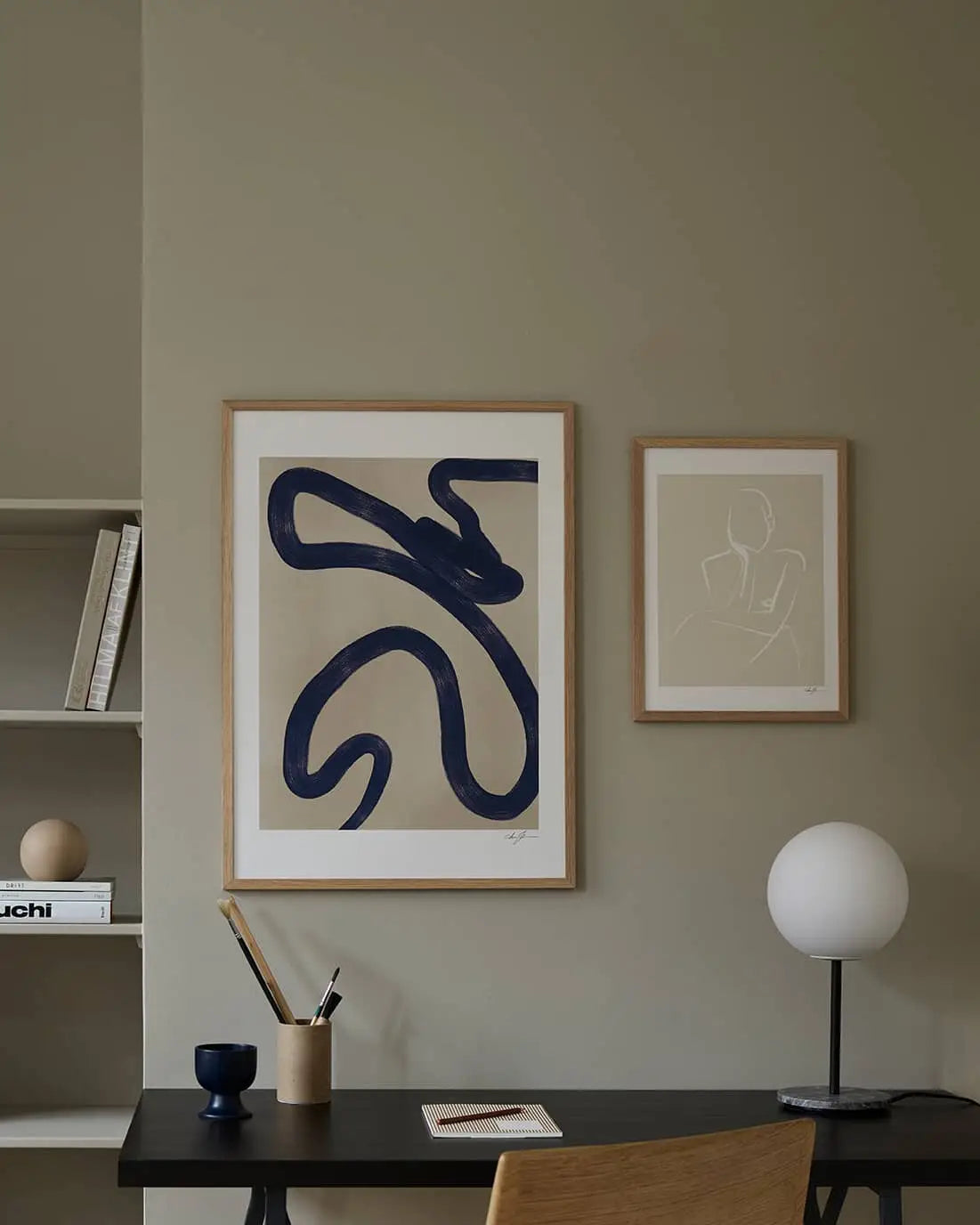 A minimalist workspace with a desk, chair, round lamp, and art supplies. Anna Johansson - Monday (30x40) by The Poster Club hangs on a beige wall above the desk; shelves with books and decor are visible to the left.