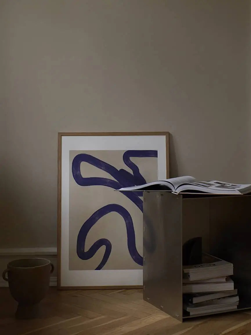 A minimalist room features The Poster Club Anna Johansson - Monday (30x40) art print with bold dark blue lines, framed and leaning against a beige wall beside stacked books, an open book on a metallic table, and a ceramic pot.