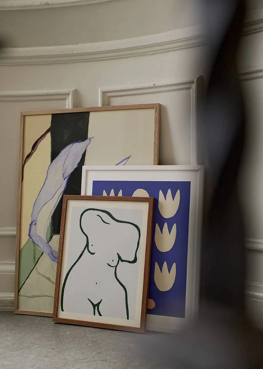 Three fine art giclée prints, including The Poster Club Anna Mörner - Statue (50x70), lean against a white paneled wall on premium art paper as a blurred figure passes by.