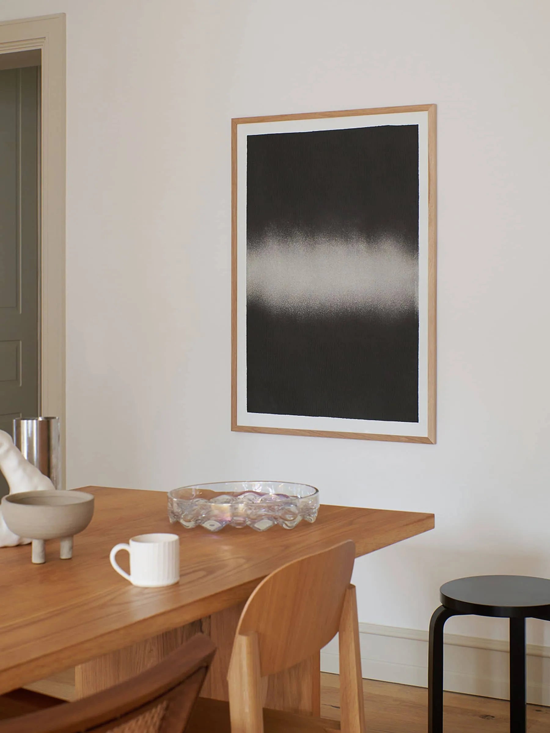 A modern dining area includes a wooden table, chairs, a white mug, glass bowl, and decor. On the wall hangs Black Interstellar (30x40) by Anne Nowak from The Poster Club—an abstract print with a black background and white horizontal blur.