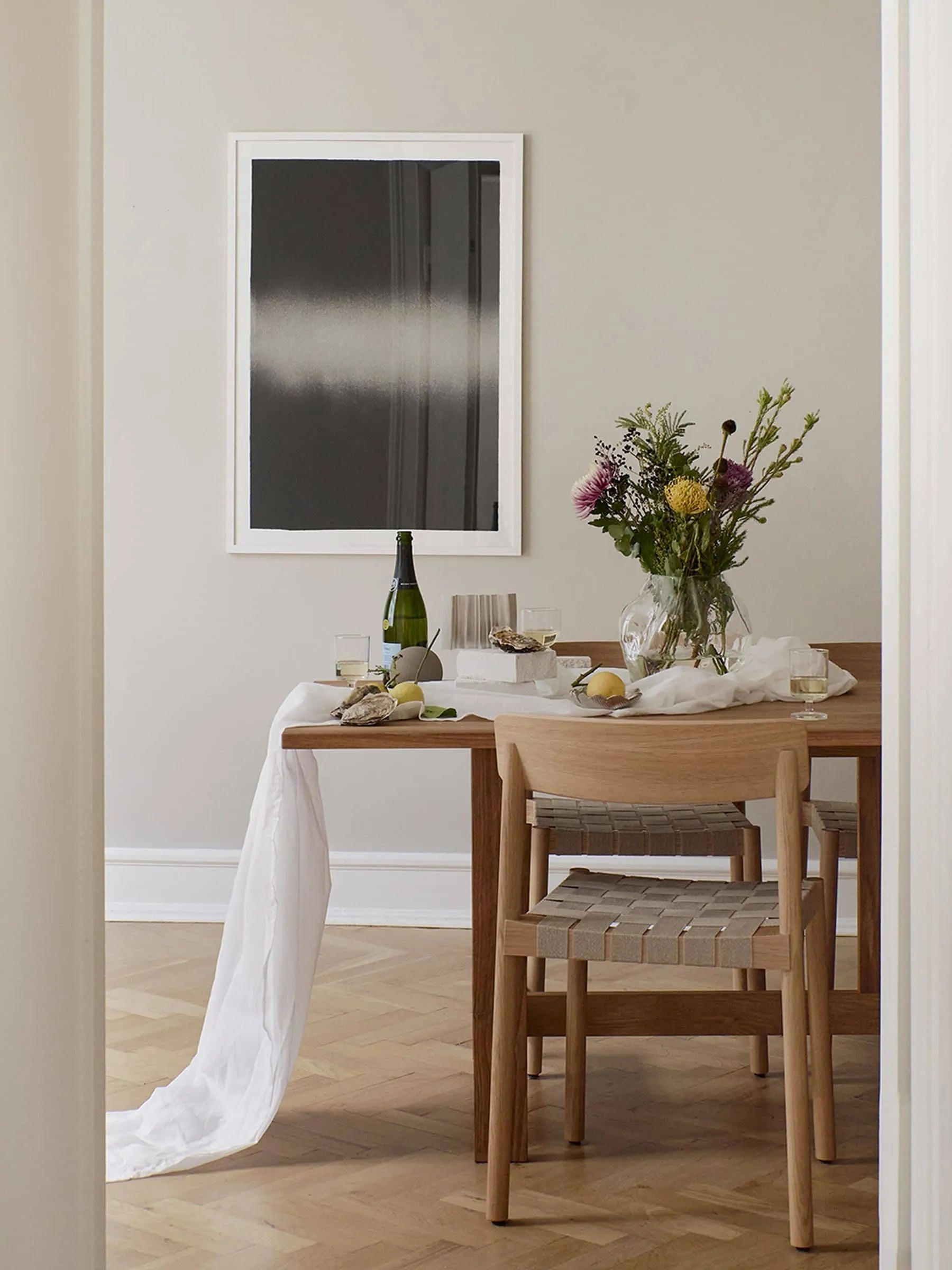 A wooden dining table with wine, glasses, flowers in a vase, food, and a white tablecloth is paired with two wooden chairs. The Poster Club’s Anne Nowak - Black Interstellar (30x40) art print completes the minimalist dining scene.