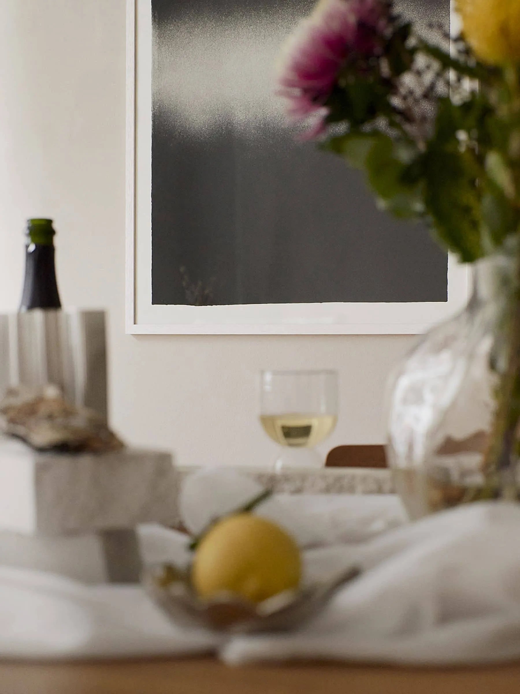 A softly focused table scene with a lemon, wine glass, bottle, and vase of flowers. On the wall hangs The Poster Club’s Anne Nowak – Black Interstellar (30x40) art print, creating a warm, inviting atmosphere.