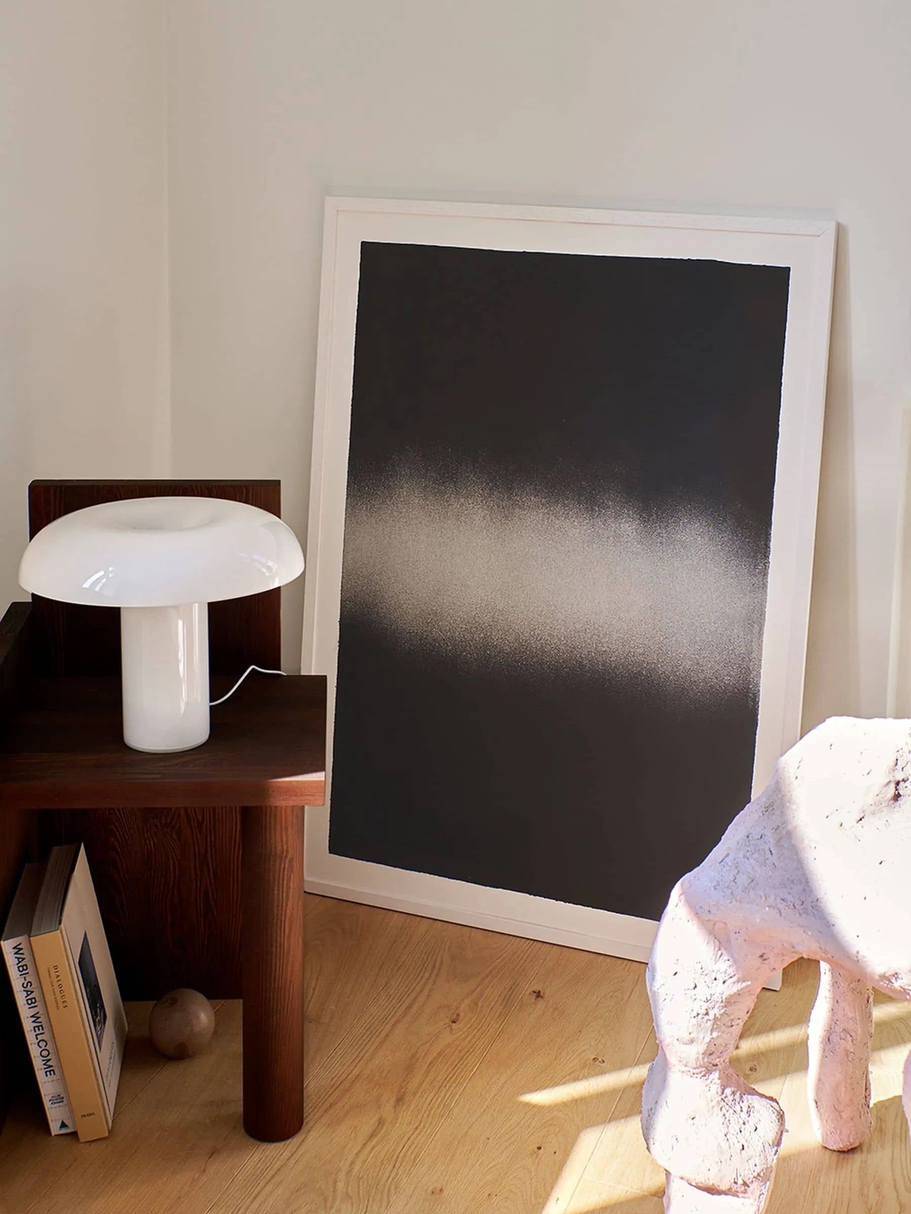 A minimalist room corner with The Poster Club’s Anne Nowak - Black Interstellar (30x40) art print leaning against the wall, a white mushroom lamp on a wooden side table, books below, and a sculptural light pink stool on wood flooring.