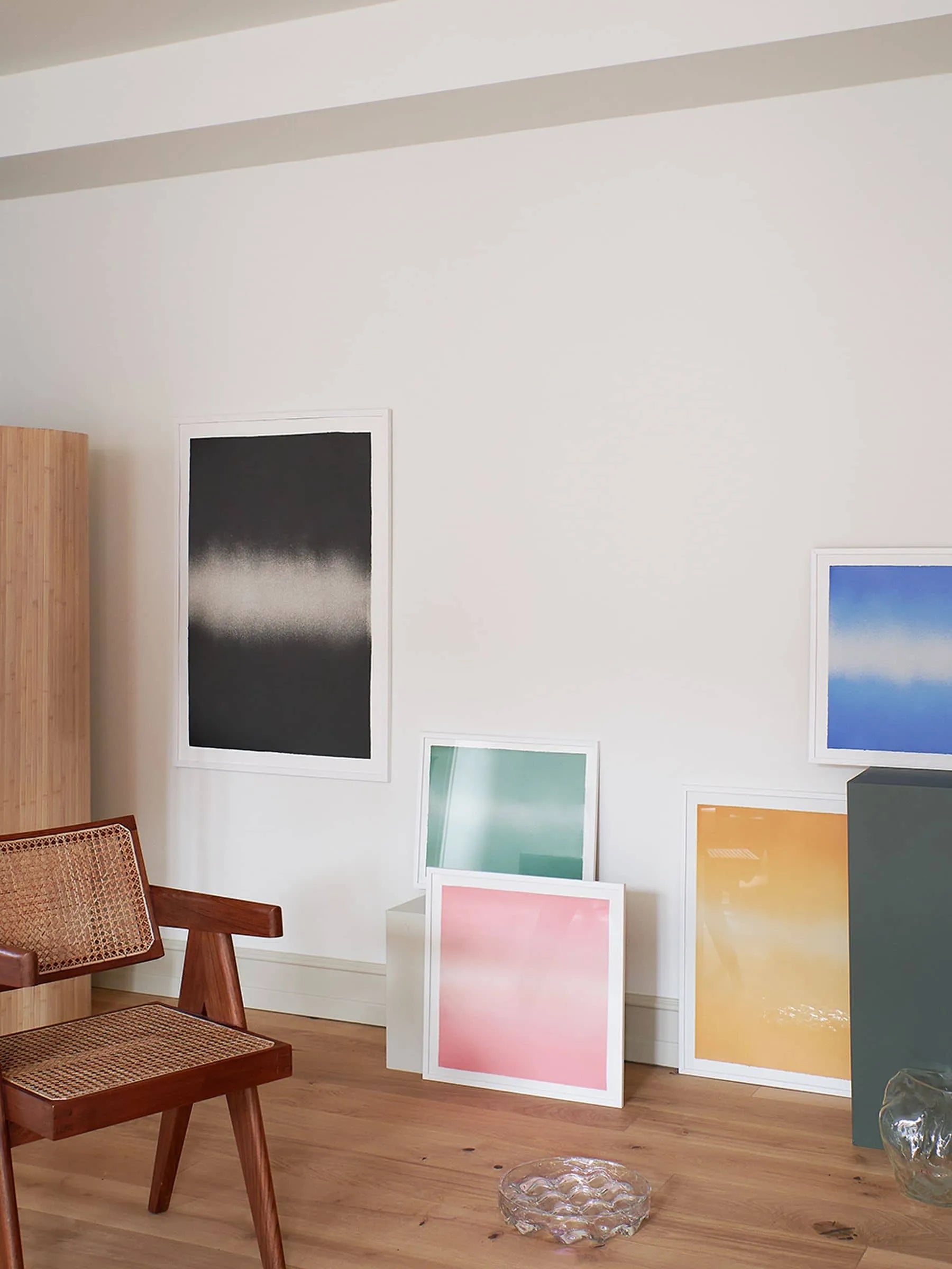 A modern room with a wooden chair, light wood flooring, and Anne Nowak’s Black Interstellar (30x40) art print by The Poster Club in a white frame displayed against a white wall.