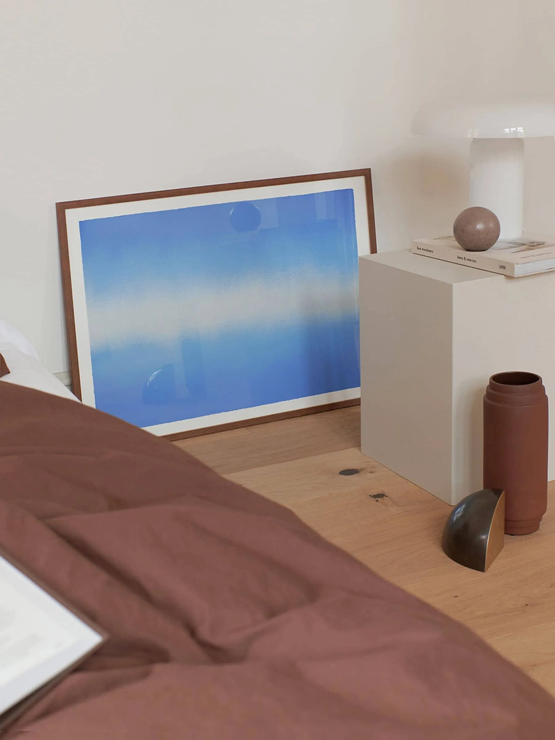 Anne Nowak’s Blue Interstellar (30x40) print from The Poster Club leans against a wall on a wooden floor beside a white cube side table with a lamp, books, and a brown sphere.