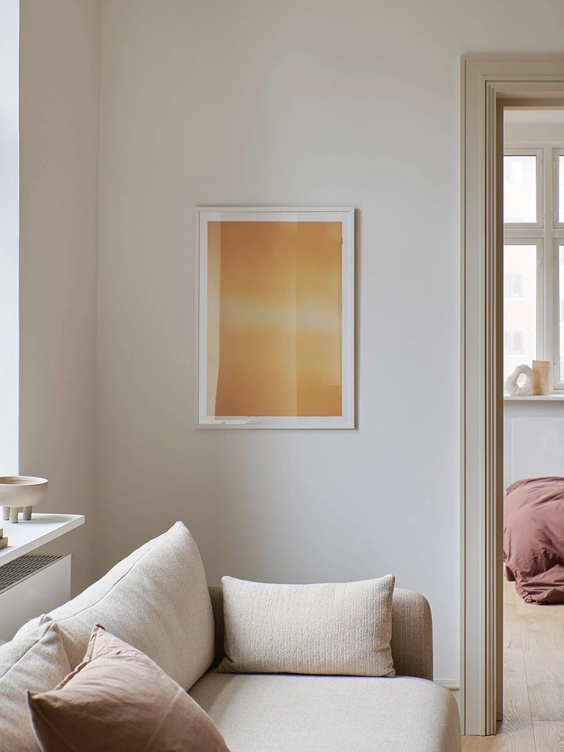 A minimal living room with a light beige sofa and cushions, a white wall displaying Anne Nowak - Yellow Interstellar (30x40) from The Poster Club, and a view into a cozy bedroom with mauve bedding and sunlight from the window.