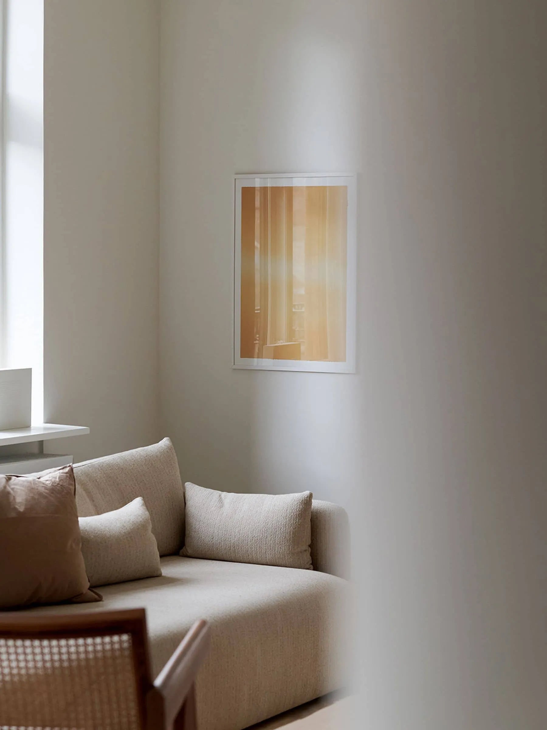 A minimalist living room features a beige sofa, cushions, wooden chair, large sunlit window, and The Poster Club’s Anne Nowak - Yellow Interstellar (30x40) art print from The Interstellar Collection simply framed on the white wall.