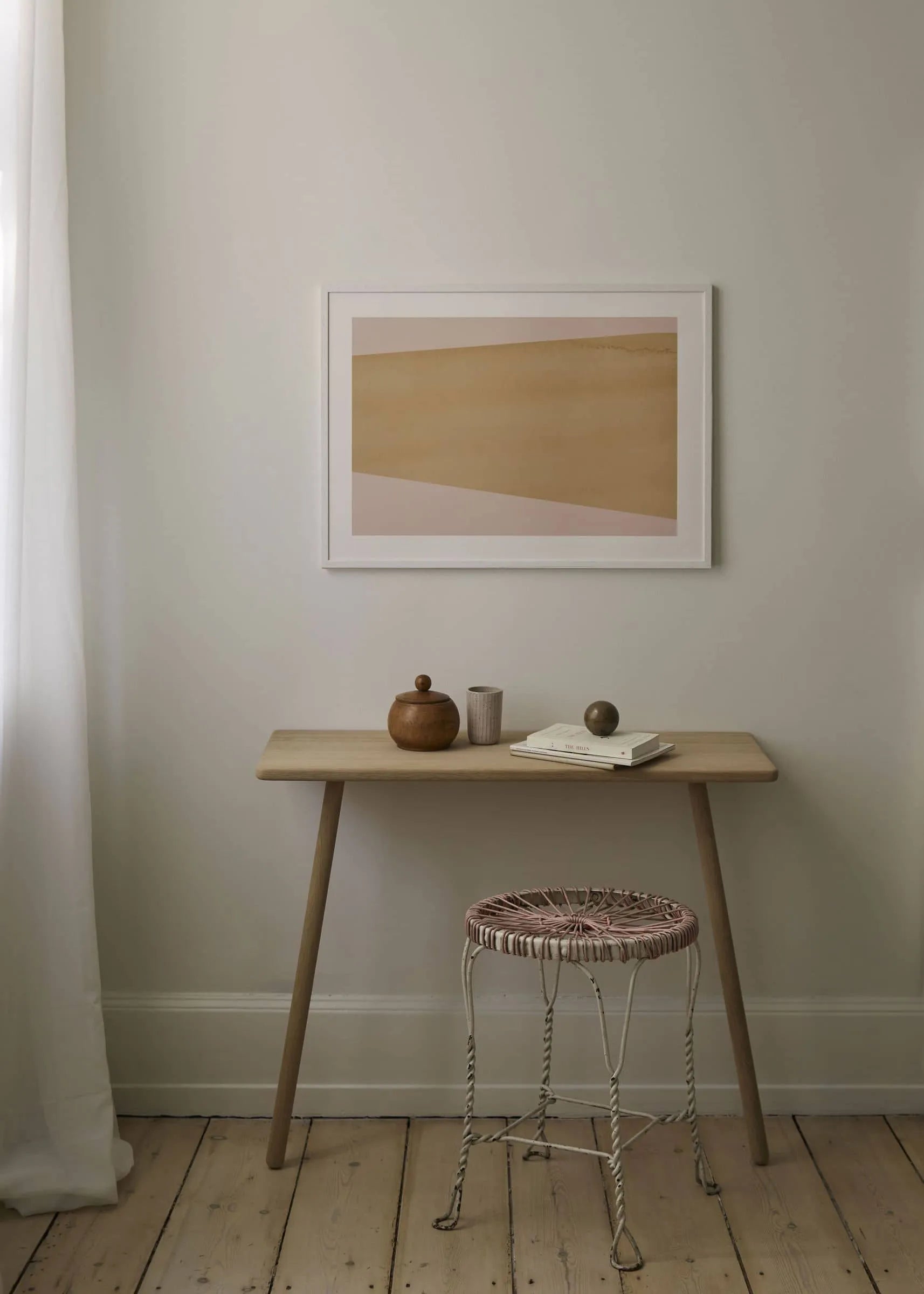 A minimalist room features a light wood desk, woven stool, neutral decor, and Atelier by Mintstudio - The E1027 Collection No. 05 (30x40) art print from The Poster Club. Soft daylight filters through white curtains onto books and beige abstract art.