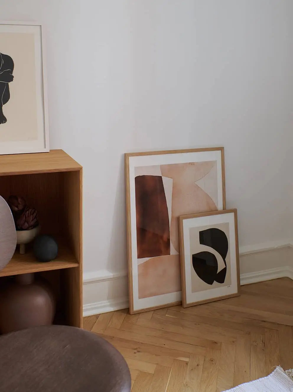 Three Berit Mogensen Lopez Bloom (50x70) art prints from The Poster Club, two leaning on a white wall and one on a wooden shelf, bring style to a cozy room corner with minimalist furniture.