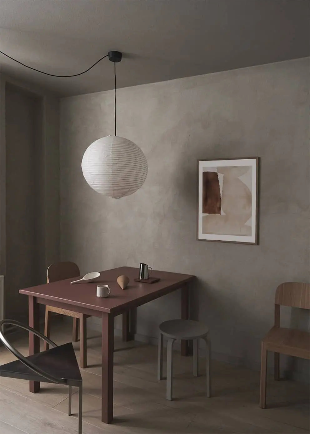 Minimalist dining area with a red table, mismatched chairs, simple pottery, a round paper pendant lamp, and The Poster Club Berit Mogensen Lopez - Bloom (50x70) art print on muted gray walls over a wooden floor.