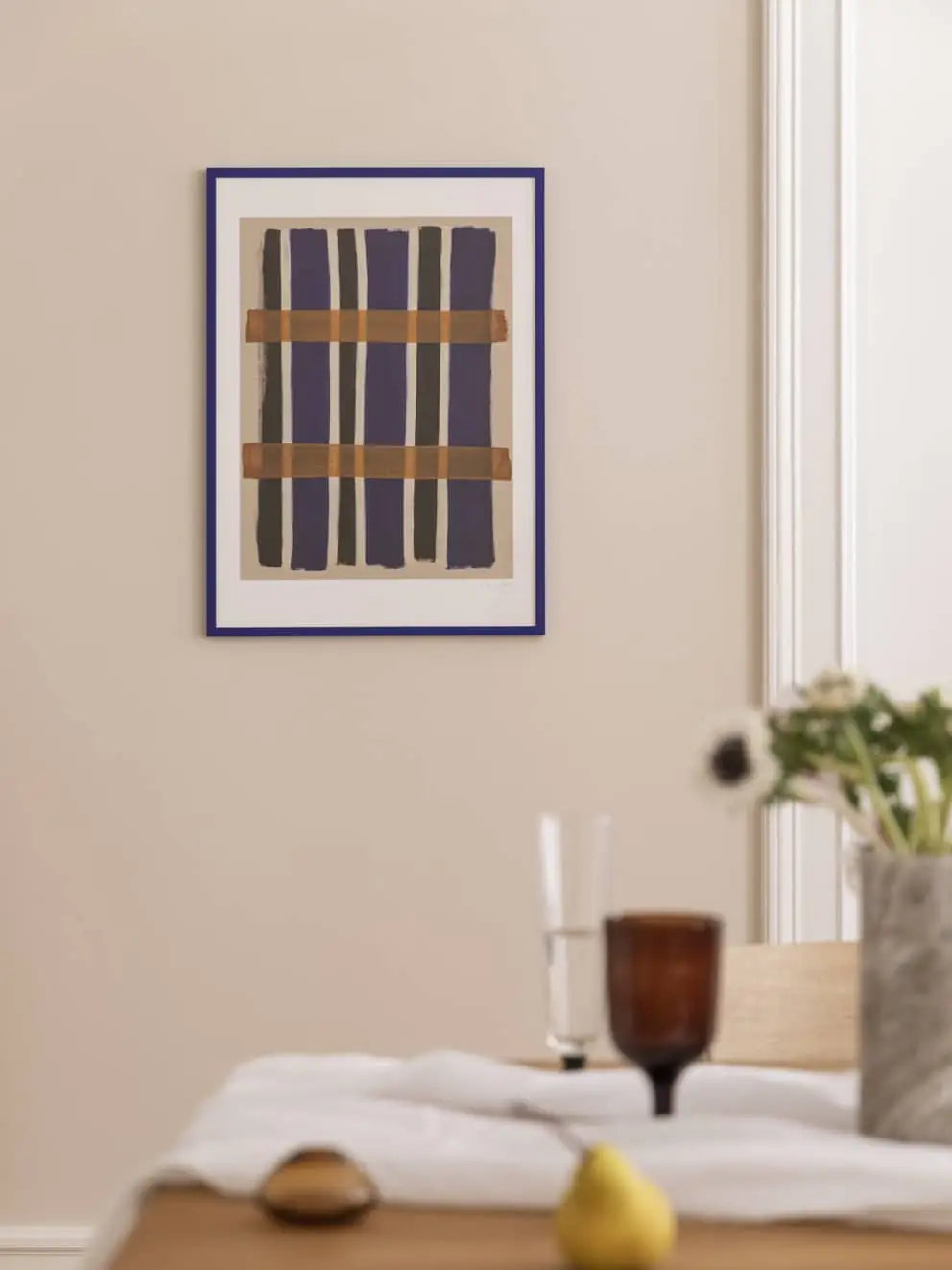 The Poster Club Berit Mogensen Lopez - Improvise (50x70) framed art print with blue and brown lines hangs on a beige wall above a table set with a glass, brown cup, white flowers in a vase, and a yellow pear.