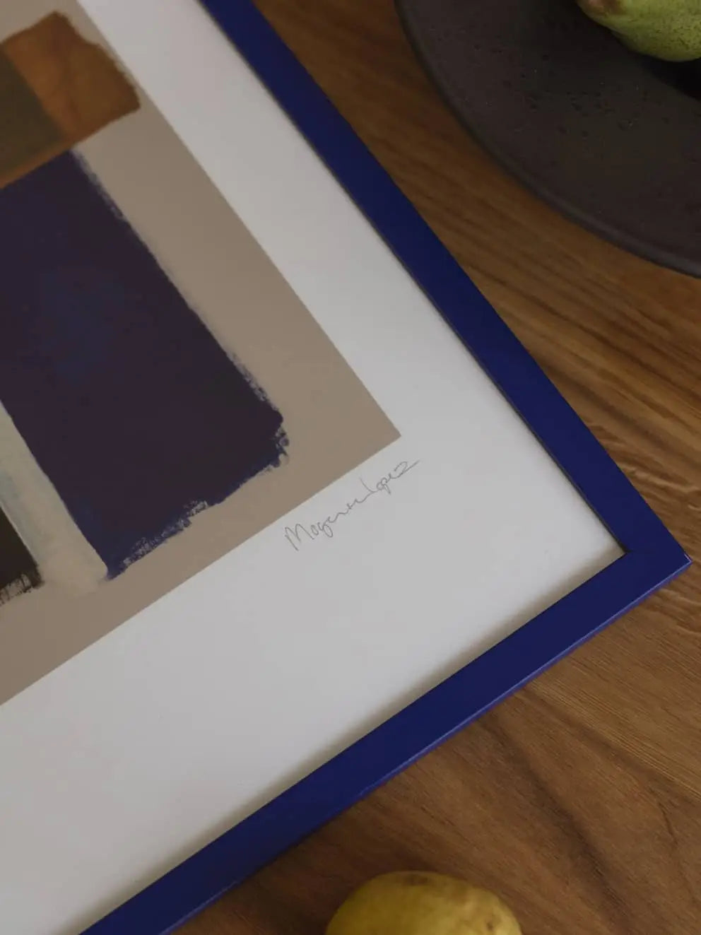 A framed Improvise (50x70) art print by Berit Mogensen Lopez from The Poster Club, with blue, brown, and beige blocks in a blue frame. The artist’s signature appears on the white border. Displayed on wood near a yellow fruit.