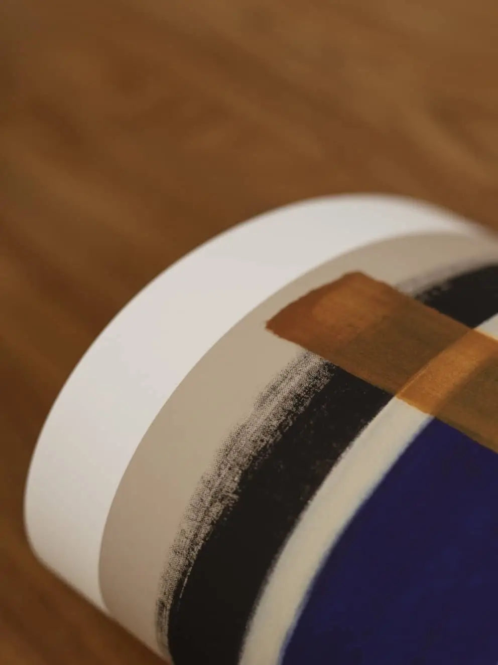 A close-up of The Poster Club Berit Mogensen Lopez - Improvise (50x70) art print, with bold black, white, and blue brushstrokes and a brown rectangle, rolled up on a wooden surface.