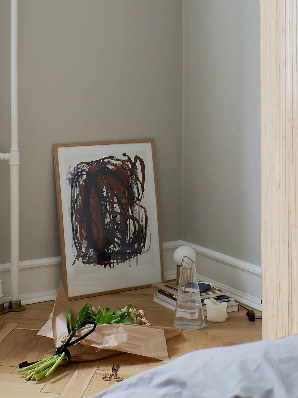 The Poster Club Berit Mogensen Lopez - Improvise (50x70) art print rests against a wall in a cozy corner, surrounded by books, a water carafe, candle, and fresh wrapped flowers on the wooden floor. Available at The Poster Club.