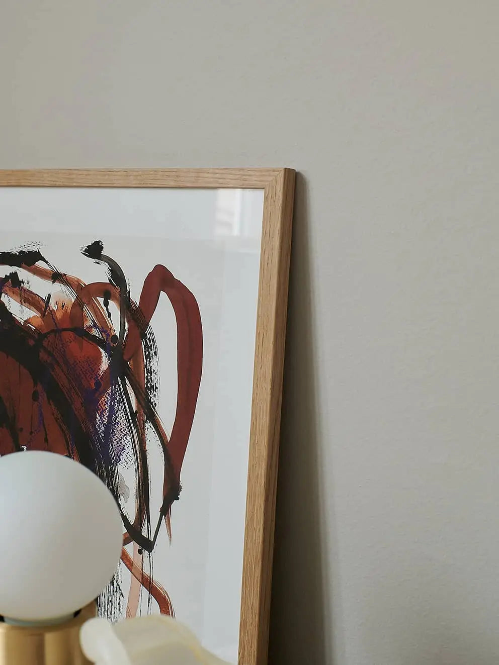 A close-up of The Poster Club Berit Mogensen Lopez - Improvise (50x70) art print, with dark red and black brushstrokes, leaning against a beige wall. A white lamp with a round bulb appears partially in the foreground.