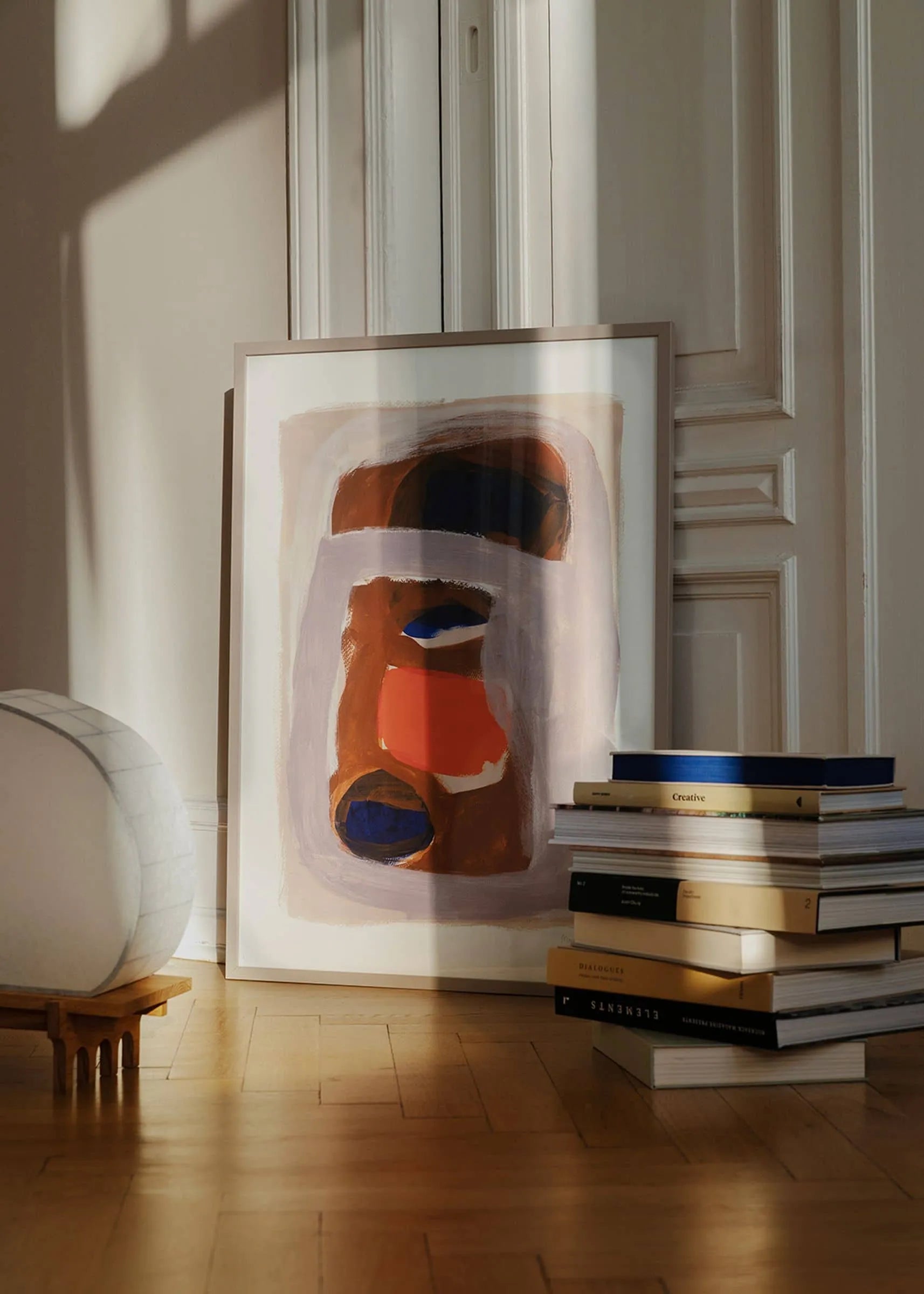 The Poster Club’s “Berit Mogensen Lopez - Outline (30x40)” framed art print leans against a white wall by a closed door, with stacks of books and a round white lamp on the sunlit wooden floor.