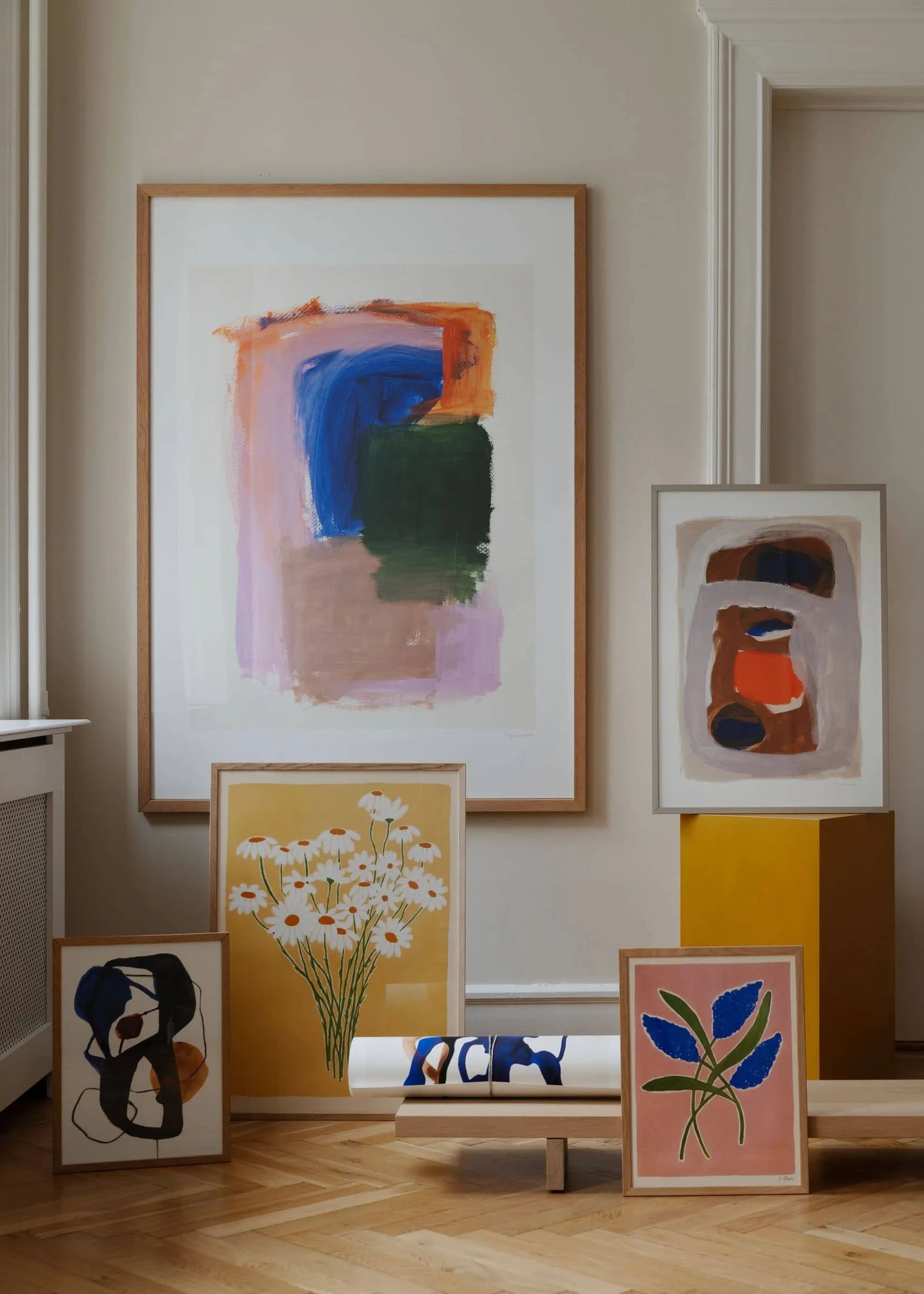 A selection of colorful abstract and floral framed paintings, including The Poster Clubs Berit Mogensen Lopez – Outline (30x40) art print, are arranged on wood flooring against a beige wall, with a large painting above and geometric collages leaning below.