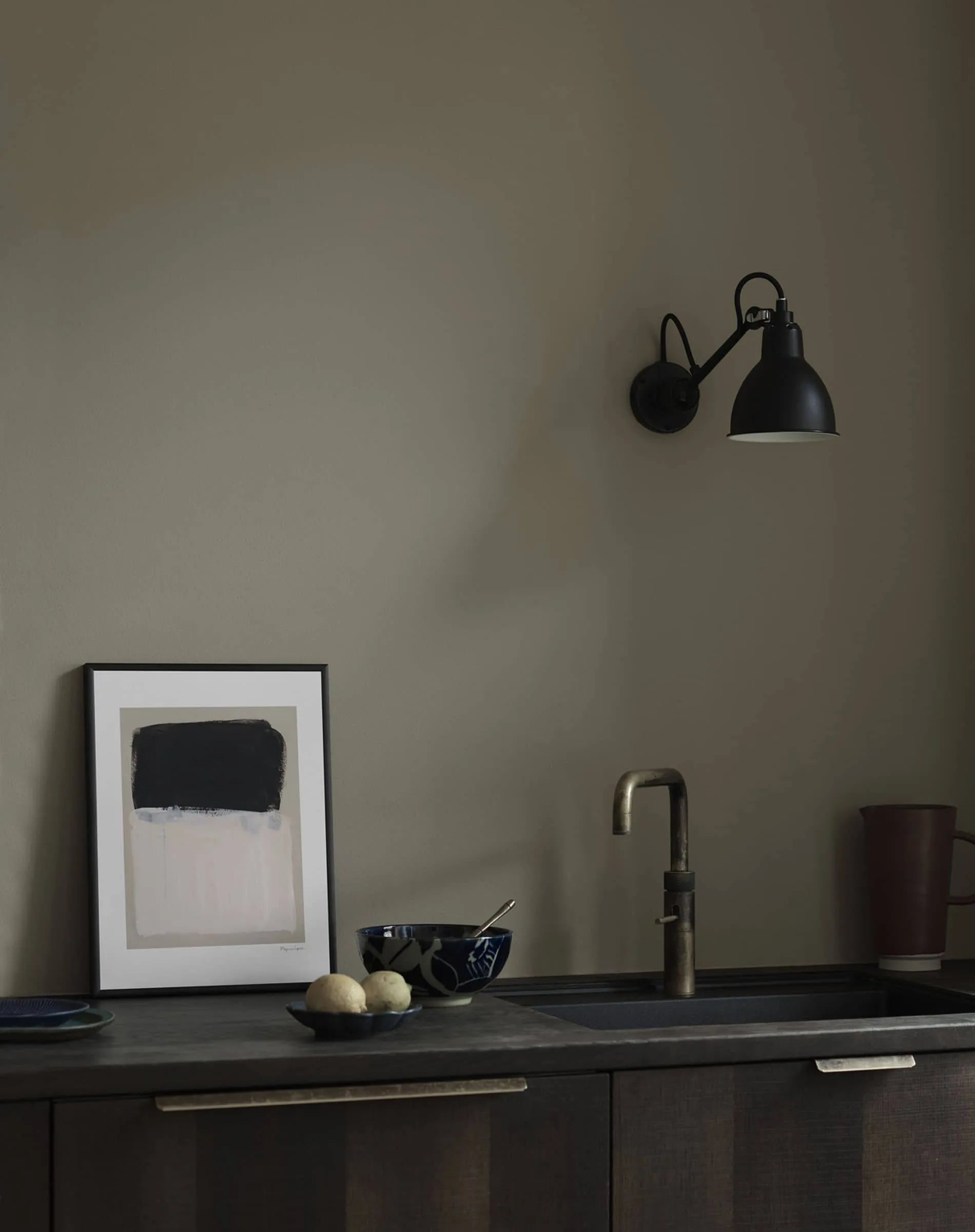 A dark, minimalist kitchen counter with a brass faucet, a bowl and spoon, two lemons, a dark cup, and Painted Surface (30x40) by Berit Mogensen Lopez from The Poster Club framed above, next to a black wall-mounted lamp.