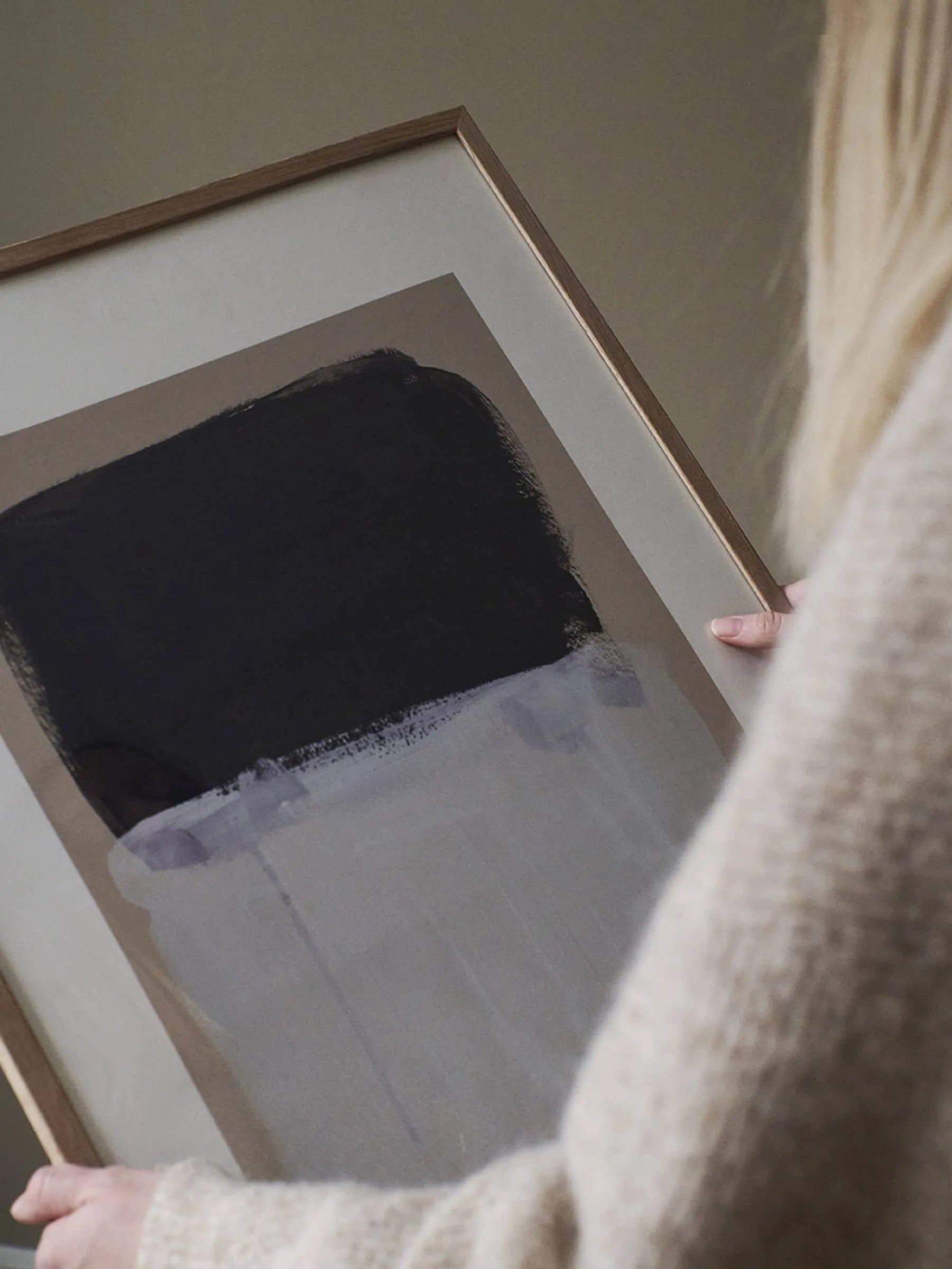 A person holds a framed Berit Mogensen Lopez - Painted Surface (30x40) art print from The Poster Club, featuring bold black, gray, and white brushstrokes against a neutral background—an elegant and modern statement piece.