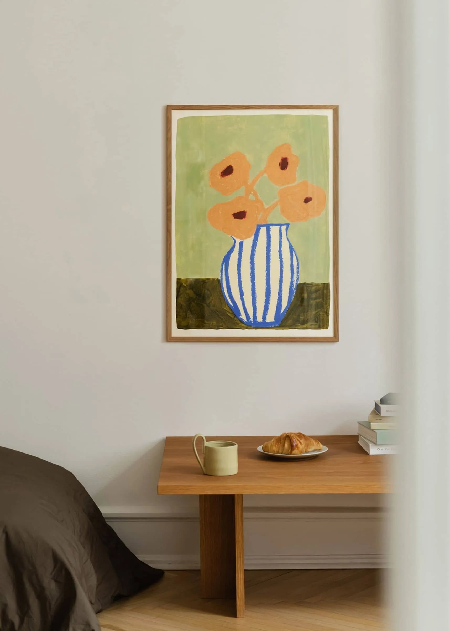 A minimal room has a wooden table with a mug, a plate with a croissant, and stacked books. On the light wall hangs The Poster Club’s Carla Llanos - Pink Flowers (30x40) fine art print above the table.