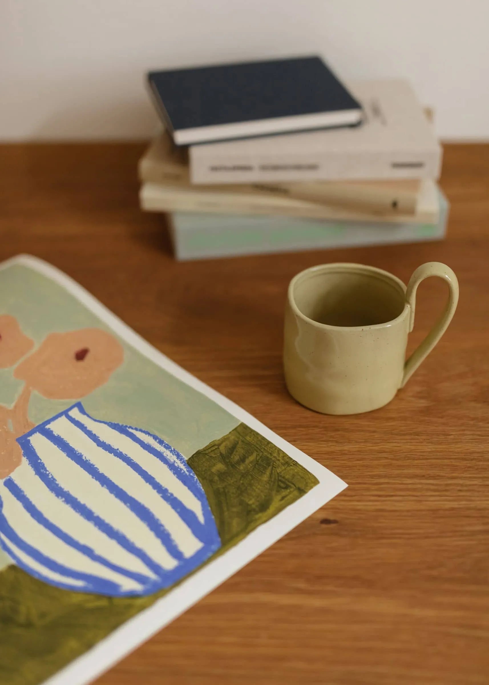 A ceramic mug sits on wood beside books. In front, The Poster Club’s Carla Llanos - Pink Flowers (30x40) artwork displays abstract floral shapes in a striped vase, resembling a fine art giclée print.