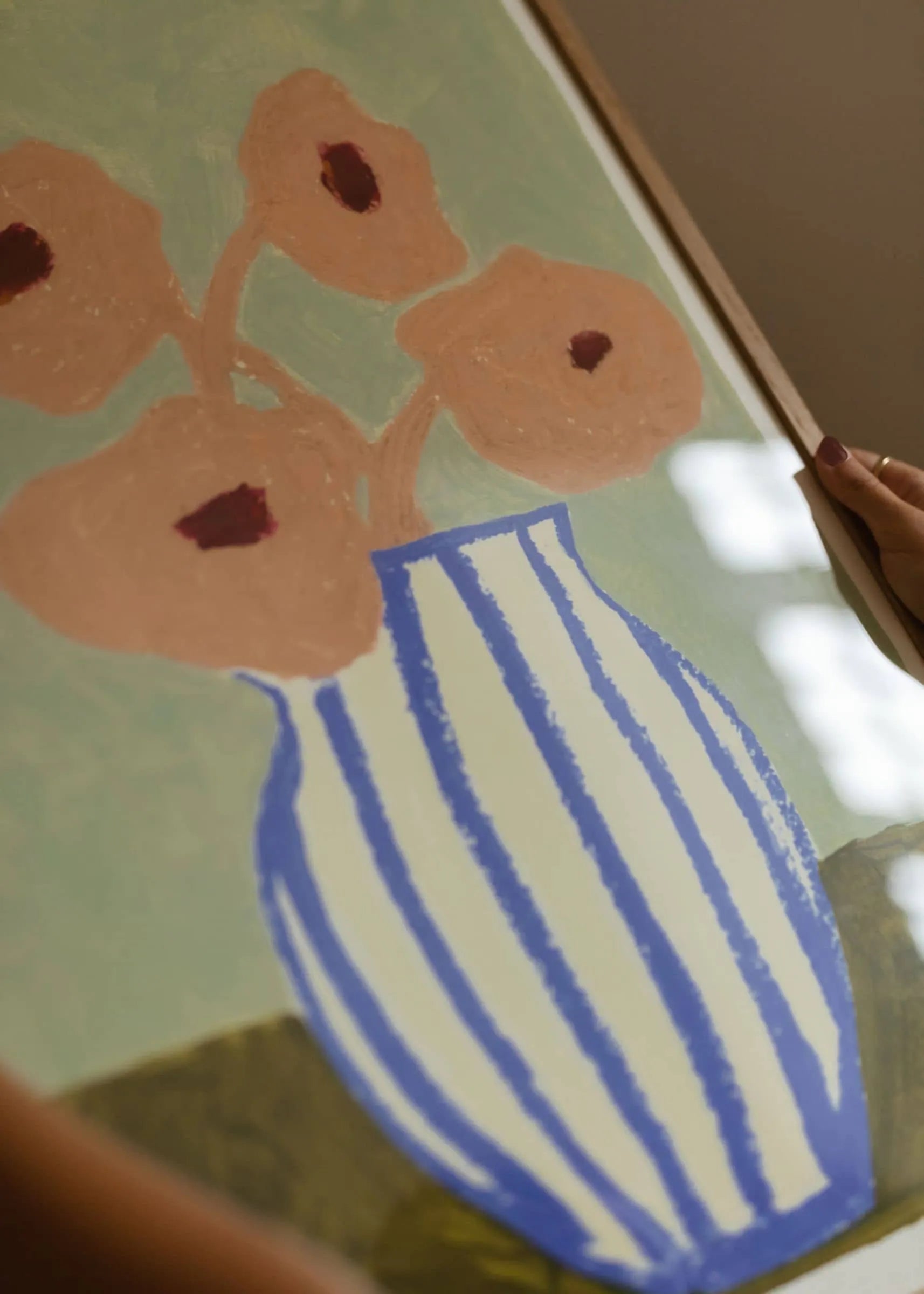 A person holds Carla Llanos - Pink Flowers (30x40) by The Poster Club, a fine art giclée print showing pink flowers with brown centers in a blue and white striped vase on a muted green abstract background.