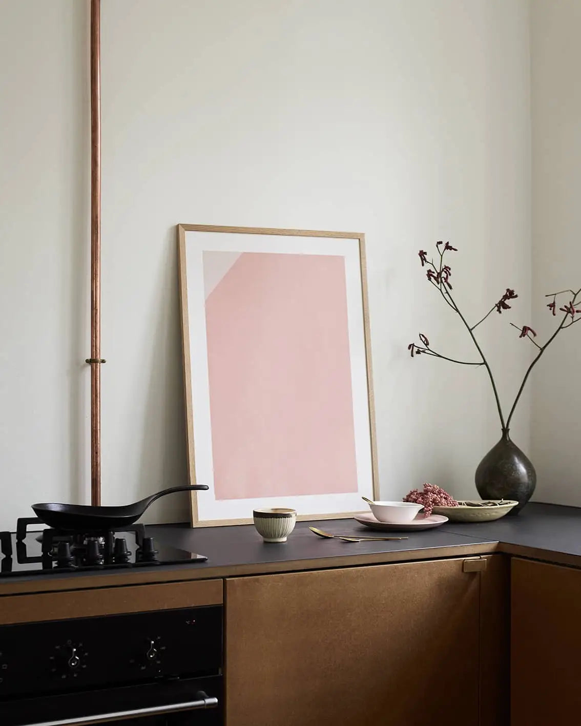 A minimalist kitchen featuring brown cabinets, a black countertop, simple dishware, a dark vase with branches, and Monday (30x40) by Anna Johansson from The Poster Club as framed abstract pink wall art.