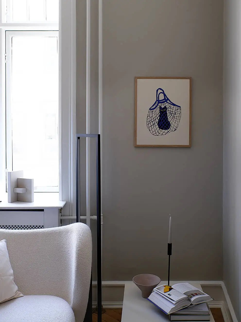 A minimalist living room features a white textured sofa, side table with candle, bowl, and open book, and The Poster Club Anna Johansson - Monday (30x40) art print on a gray wall. Sunlight streams in through a large window.