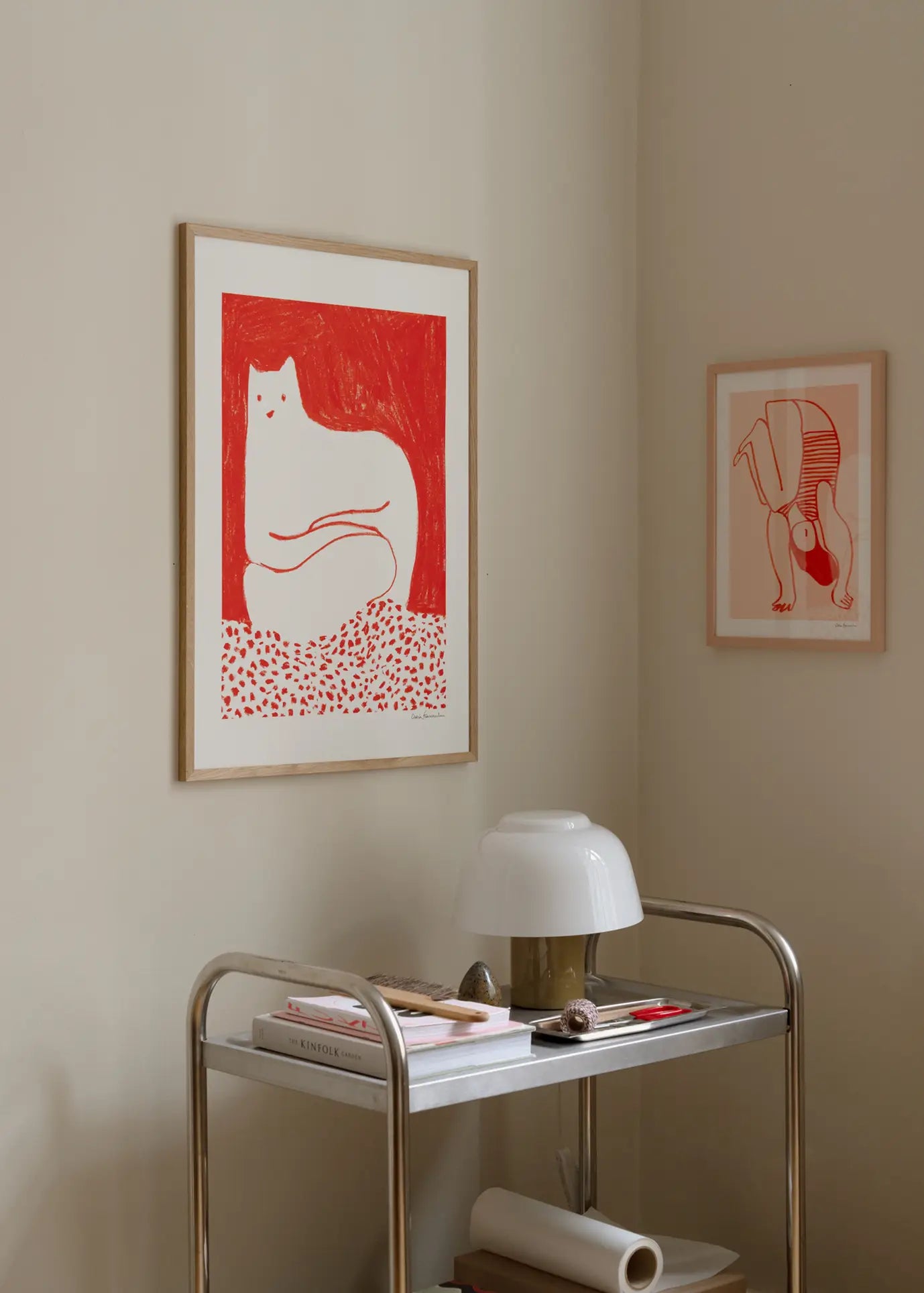 A small metal cart with books and a white lamp stands against a beige wall, decorated with two framed red and white Giclée prints—one is The Poster Club Cinzia Franceschini - Cat In Red (30x40) by The Poster Club.