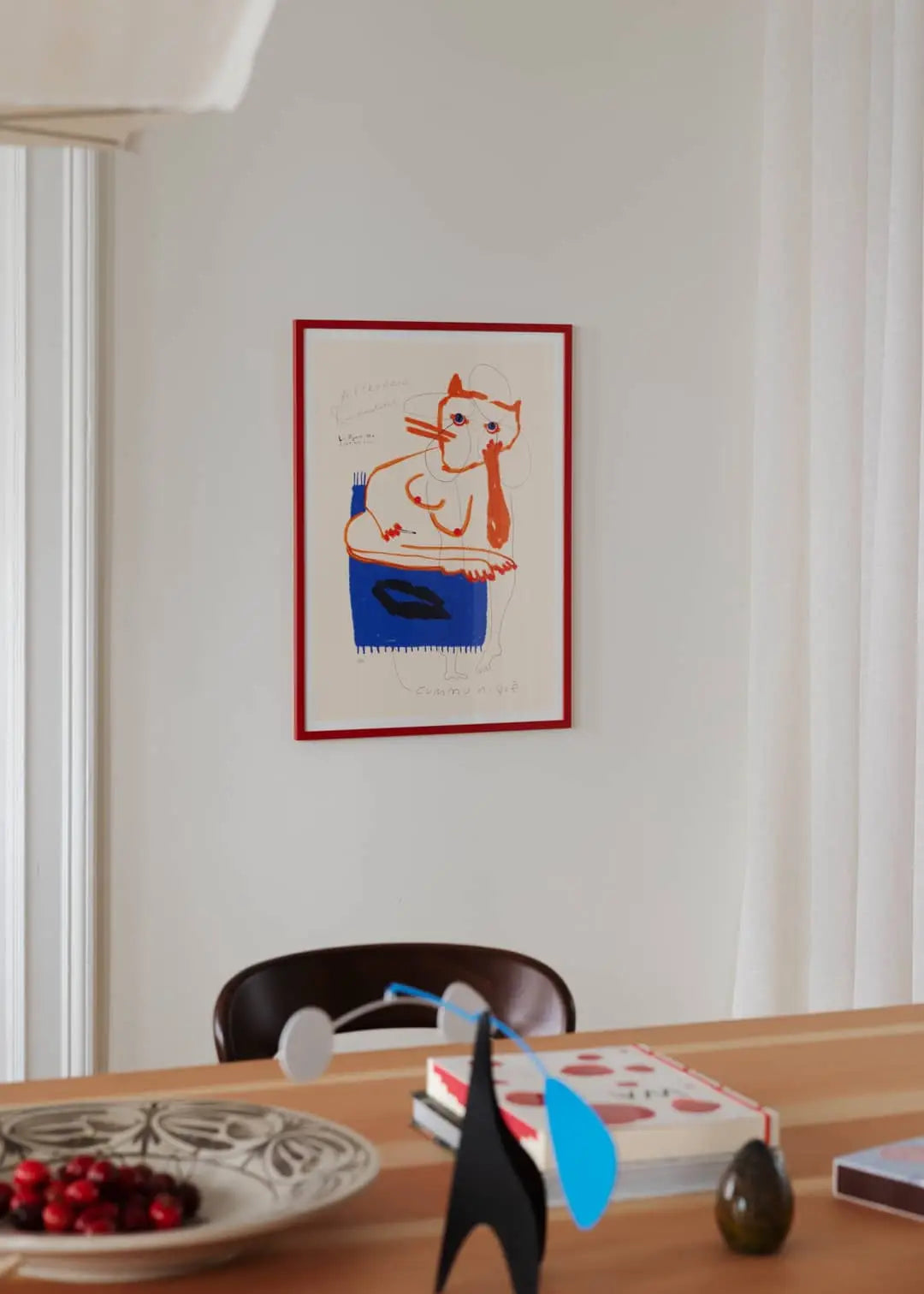 The Poster Club Das Rotes Rabbit - Afternoon Communiqué (50x70) fine art print by The Poster Club hangs above a wooden table with books, vase, blue sculpture, and cherries. A brown chair completes this stylish wall decor.