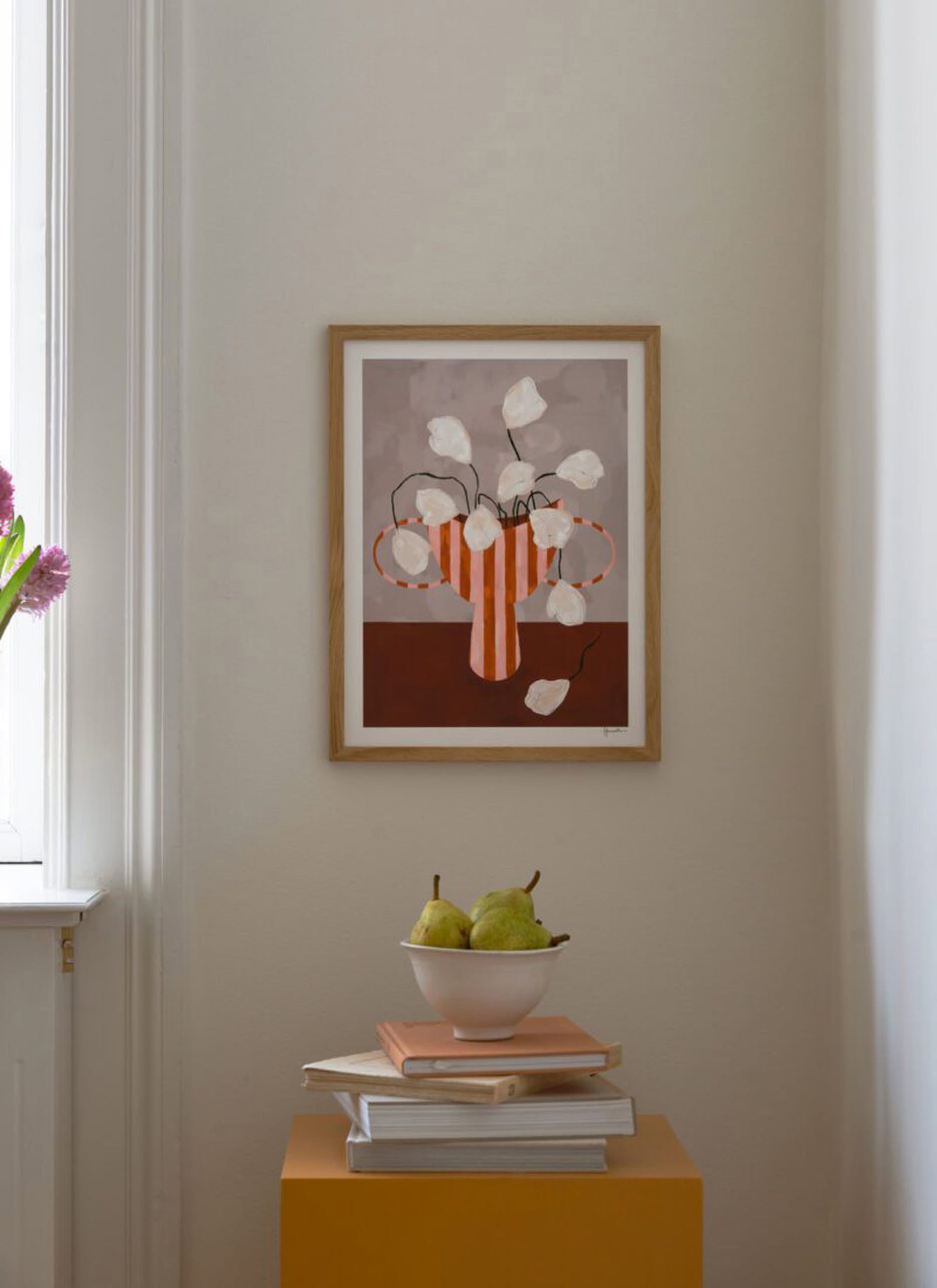 The Poster Club’s Frankie Penwill - White Flowers in Striped Vase (30x40) poster is displayed on a cream wall above a yellow table with books, a plate, and two pears, bathed in natural light from a window on the left.