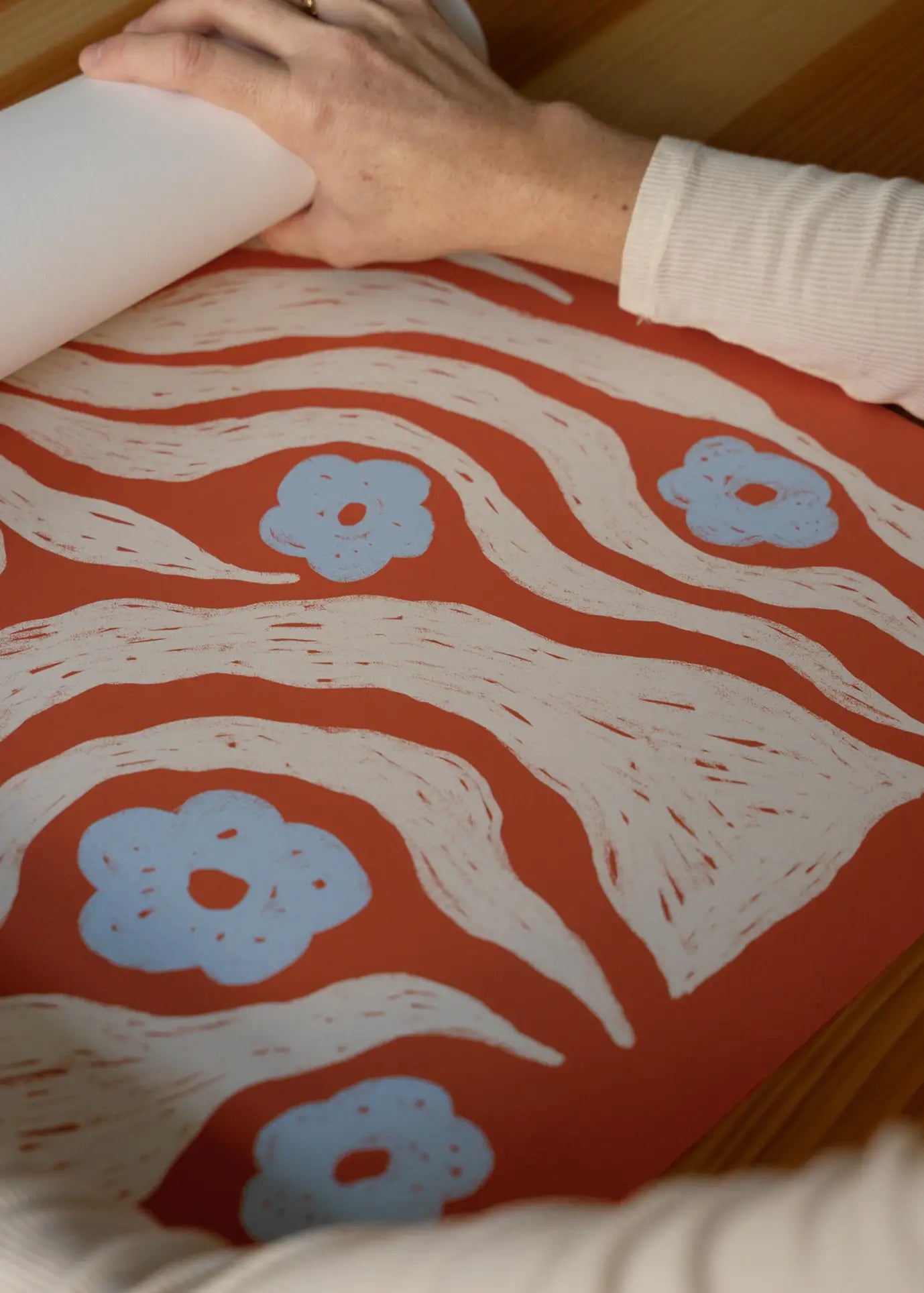 A person unrolls The Poster Club Hanna KL - Misty (50x70) art print, featuring a red background with white waves and blue flowers, on a wooden surface.