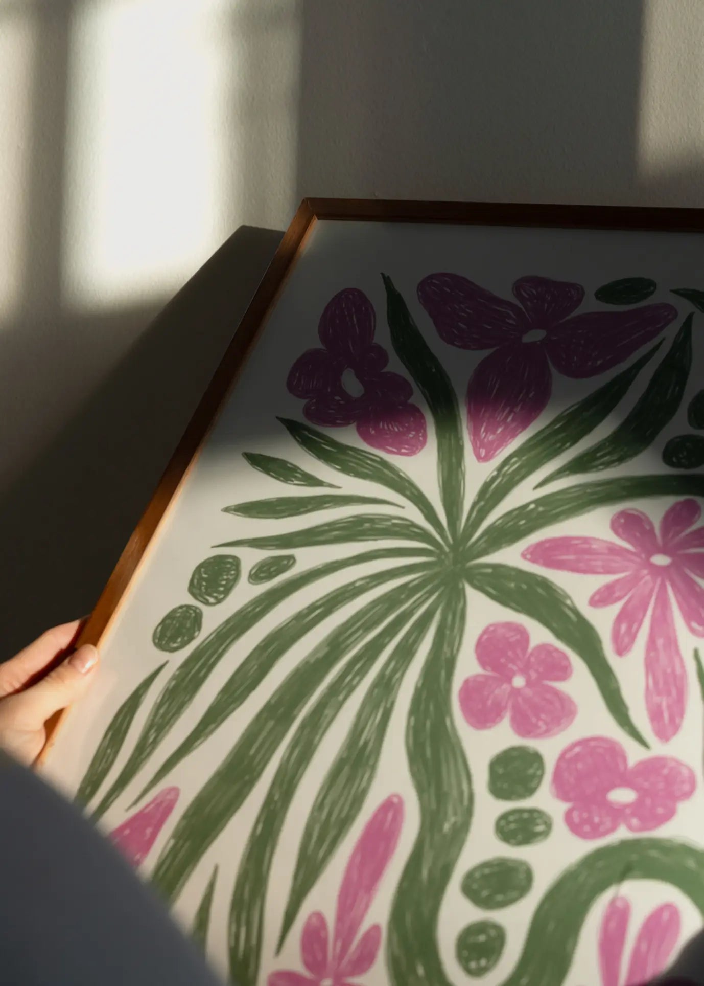 A hand holds The Poster Club Hanna KL - Wonder (50x70) art print, displaying green leaves and pink flowers with soft sunlight casting shadows behind—a tranquil piece from The Poster Club.