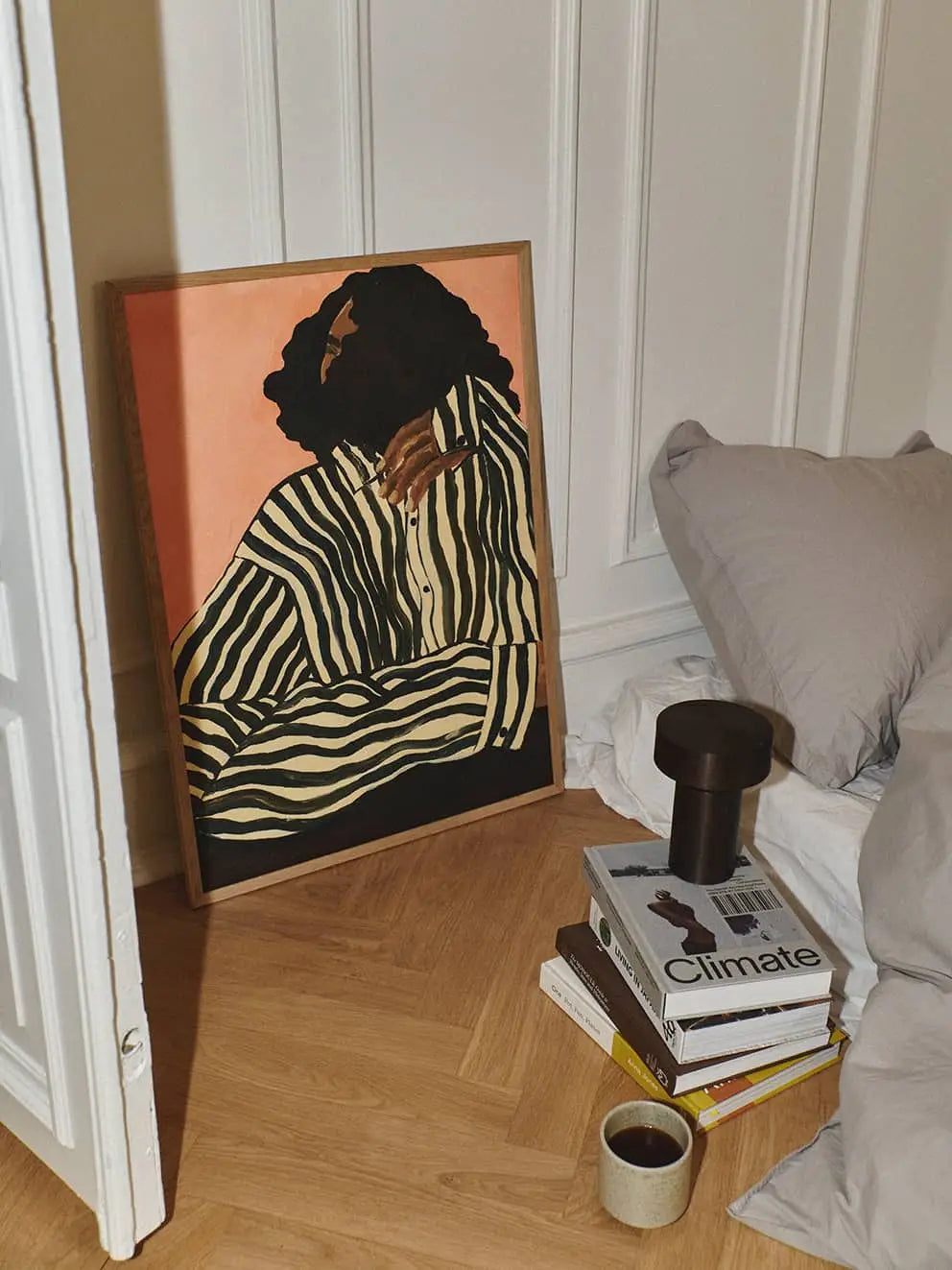 The Poster Club Anna Johansson - Monday (30x40) art print leans against a wall on a wooden floor, with stacked books, a black lamp, and a coffee cup beside a bed with gray bedding.