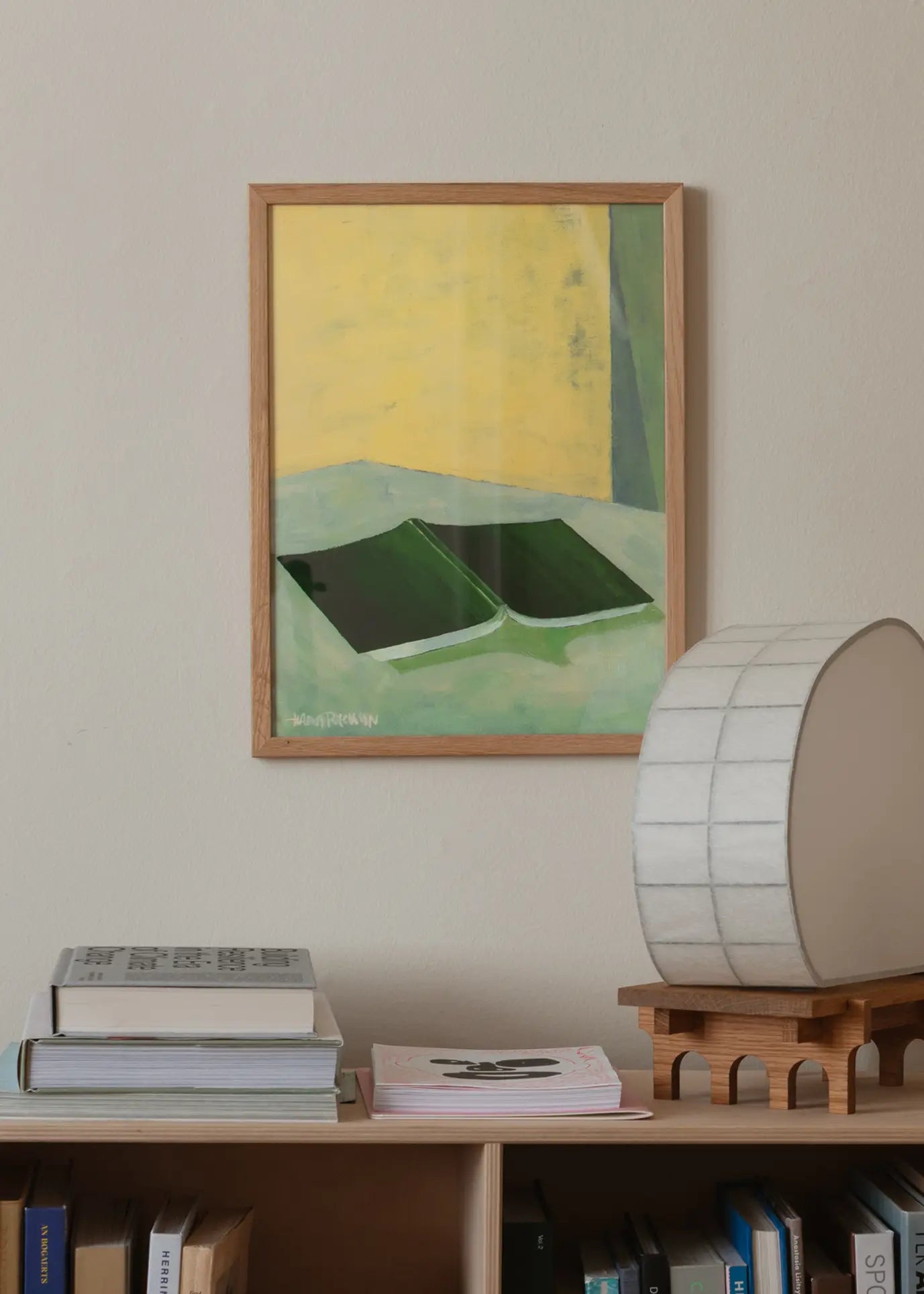 The Poster Club Hanna Peterson - The Book (30x40) fine art print features two green books on a yellow and green background, framed and displayed above a wooden shelf with books and an oval-shaped white lamp.