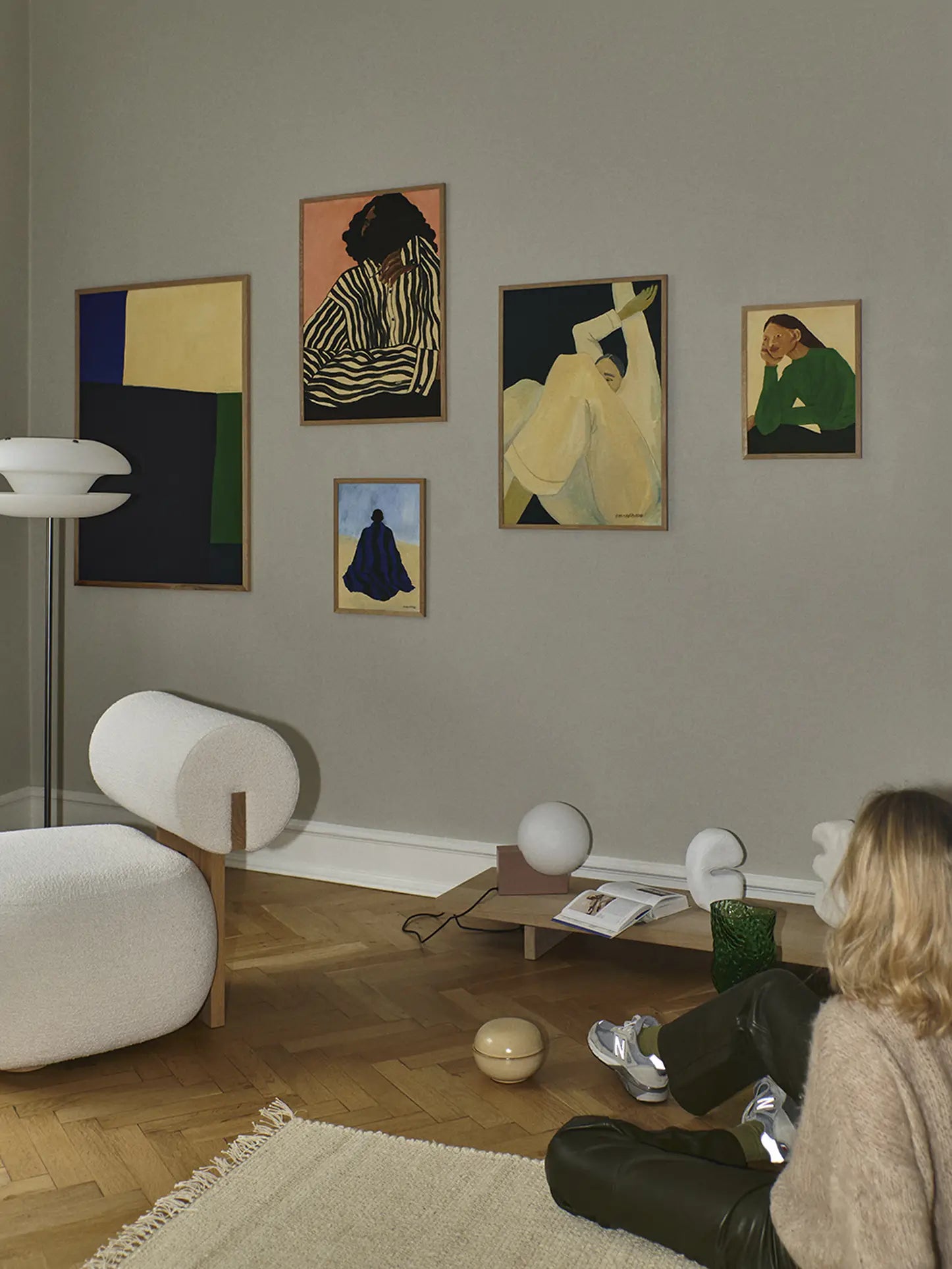 A woman sits on a beige rug in a modern living room with wooden floors. Above a white chair, the wall displays The Poster Club Hanna Peterson - The Book (50x70) fine art print in a wooden frame, surrounded by minimalist decor.