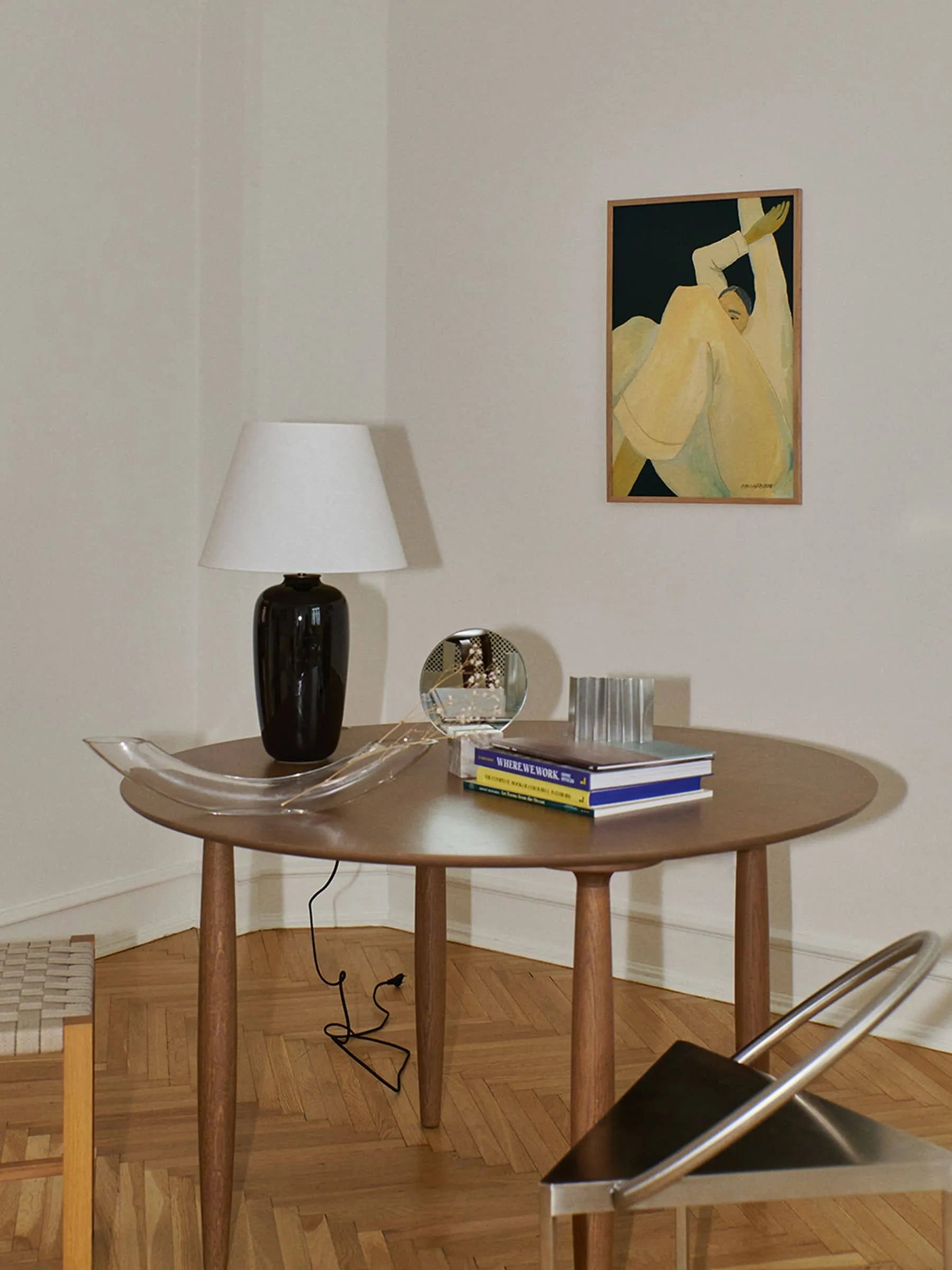 A round wooden table with a black lamp, glass tray, books, and a glass orb sits on herringbone floors. Nearby are a woven stool and metal chair; on the white wall hangs The Poster Clubs Hanna Peterson - The Dream (30x40) art print.