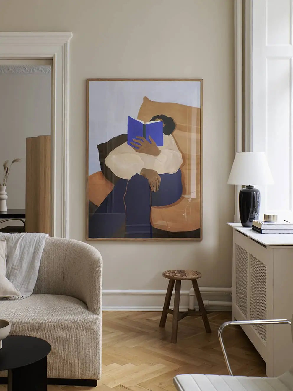 A modern living room in neutral tones features The Poster Club Anna Johansson - Monday (30x40) art print above a small wooden stool, next to a light-colored sofa and lamp. The artwork shows a person reading a blue book.
