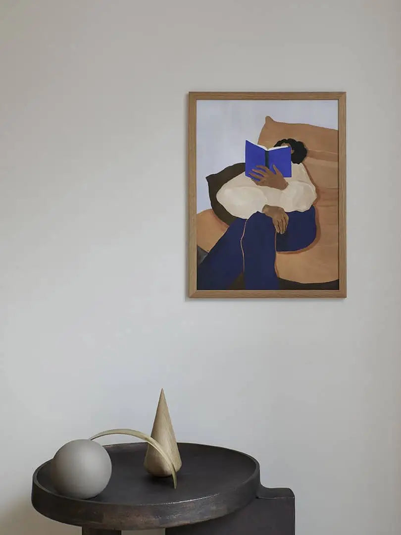 The Poster Club Anna Johansson - Monday (30x40) art print by The Poster Club depicts a person on a sofa with an open blue book covering their face, above a table with geometric decor, set against a plain white wall.