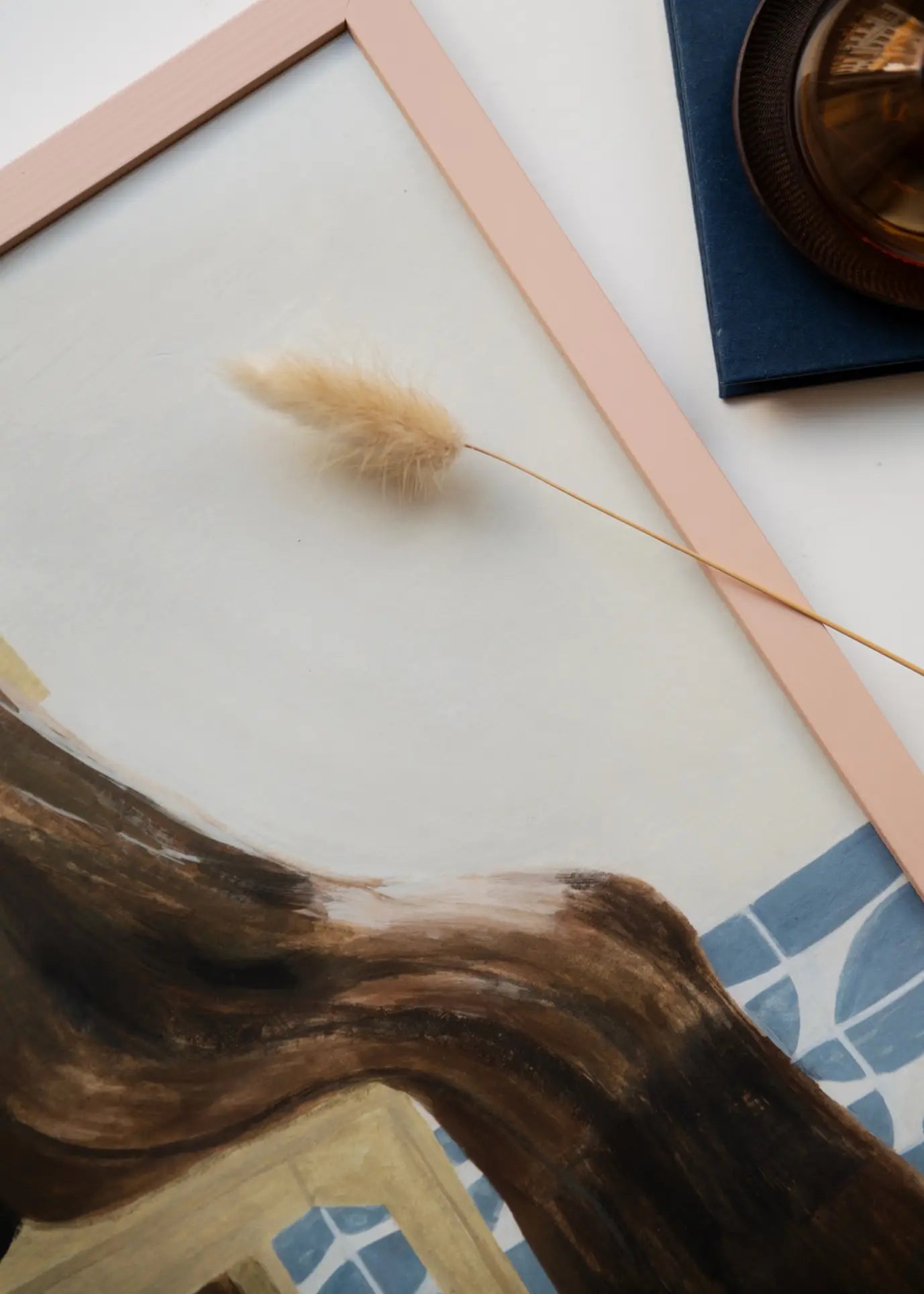 A close-up of The Poster Club’s Isabelle Vandeplassche - Chair With Blanket (70x100) art print shows soft pink edges, a figure with long brown hair, blue tile details, and nearby dried grass with a brown glass object.