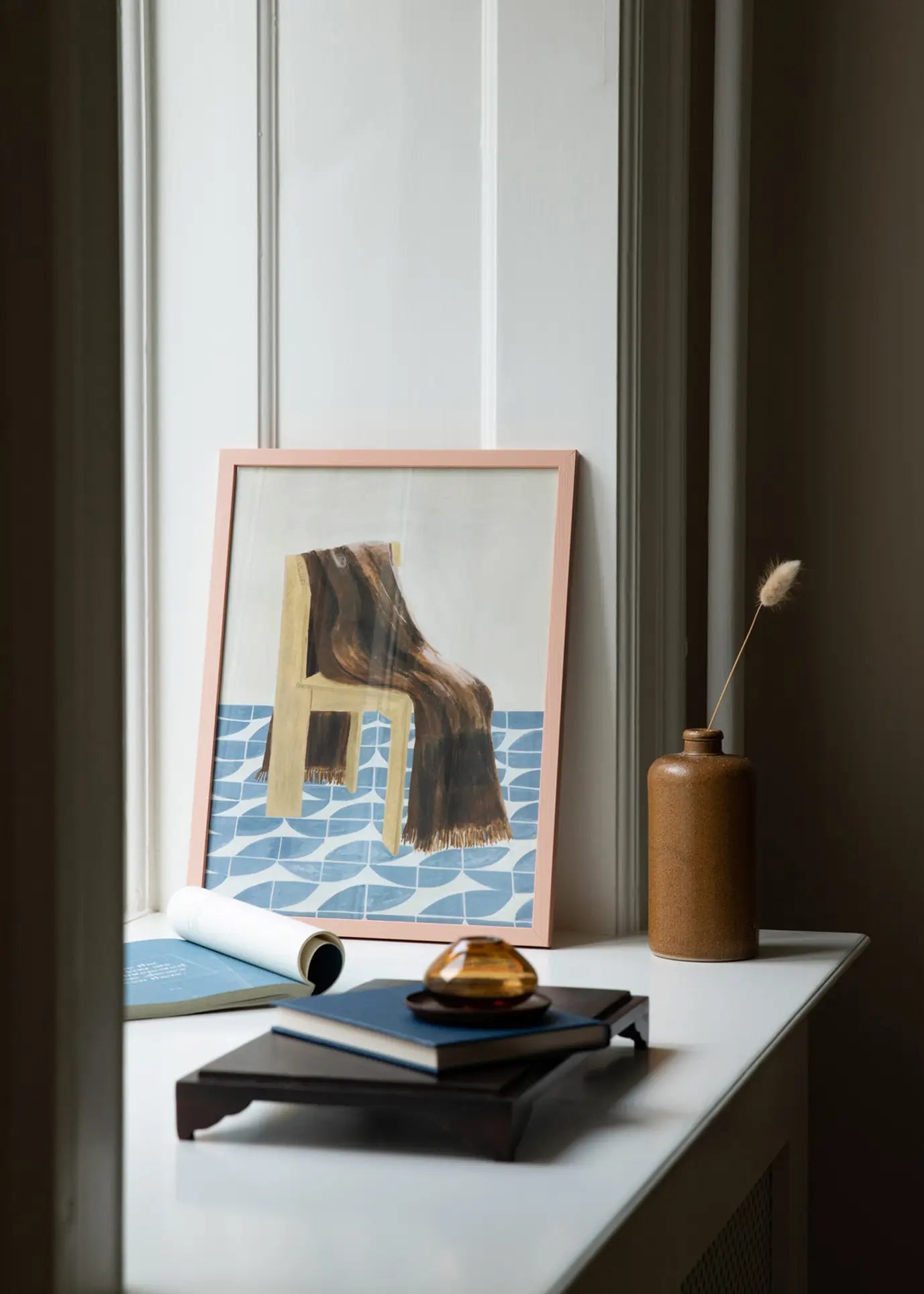 The Poster Club’s Isabelle Vandeplassche - Chair With Blanket (70x100) art print features a chair draped with a blanket by a window, next to a brown vase with dried grass, books, a magazine, and an amber glass dish.