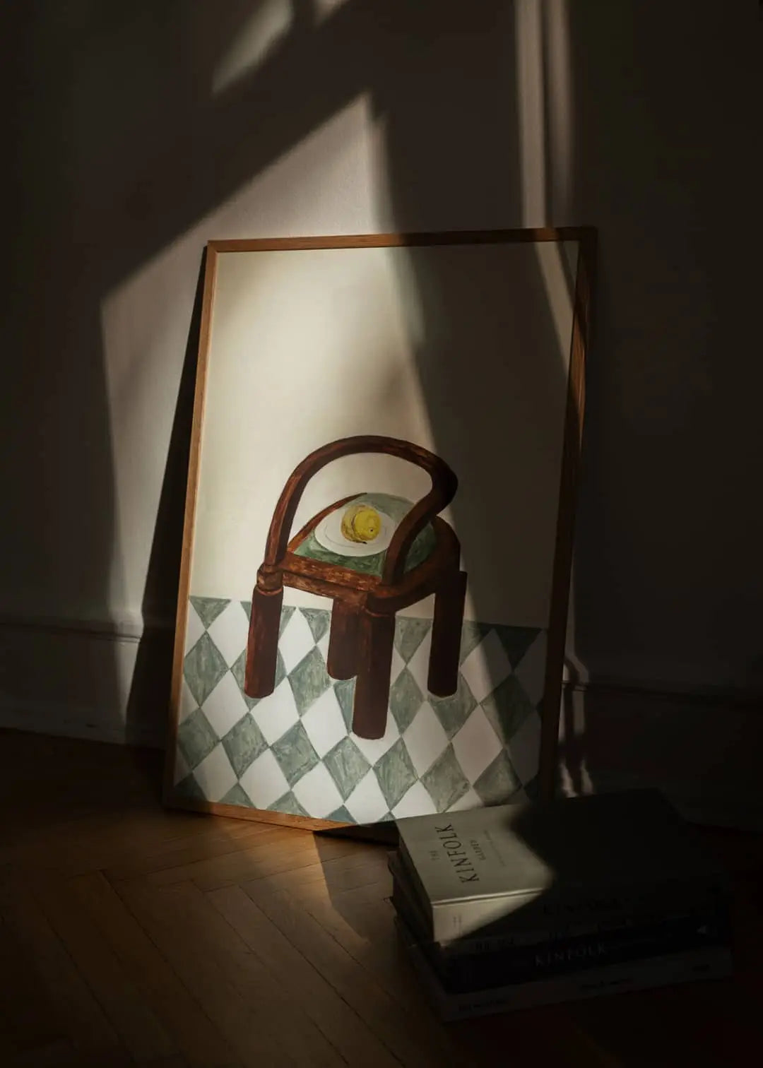 The Poster Club’s Bloom (50x70) by Berit Mogensen Lopez depicts a wooden chair with a cushion and lemon on a checkered floor in soft sunlight, books nearby—a refined framed art print from The Poster Club.