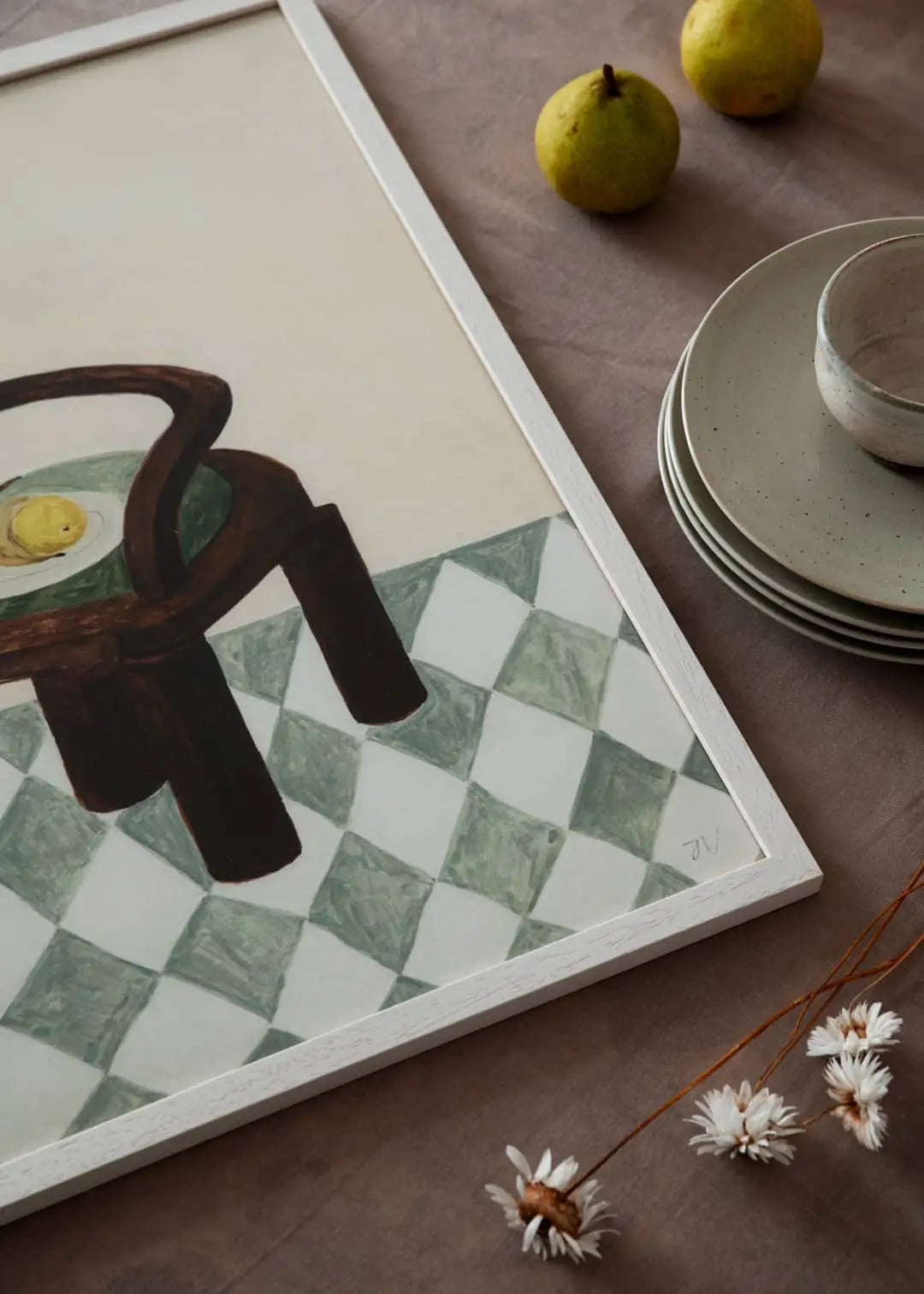 The Poster Club’s Berit Mogensen Lopez - Bloom (50x70) art print features a chair and lemon on a checkered floor, displayed framed on a brown table with stacked plates, a cup, two pears, and dried white flowers.